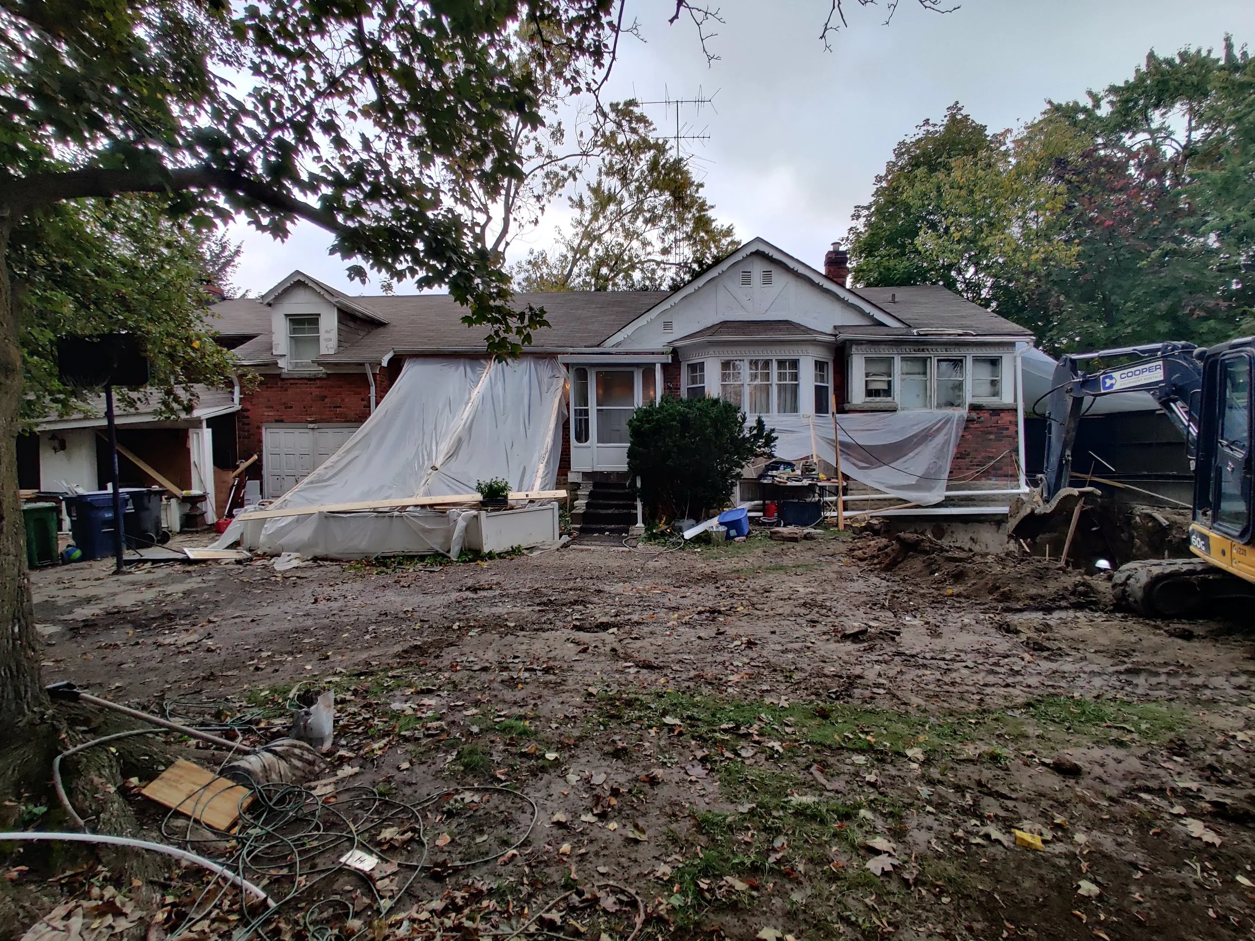 A two-story brick house with a covered porch, surrounded by trees with green leaves, and a construction site in front with a tarp, tools, and equipment, possibly undergoing renovation or repair.