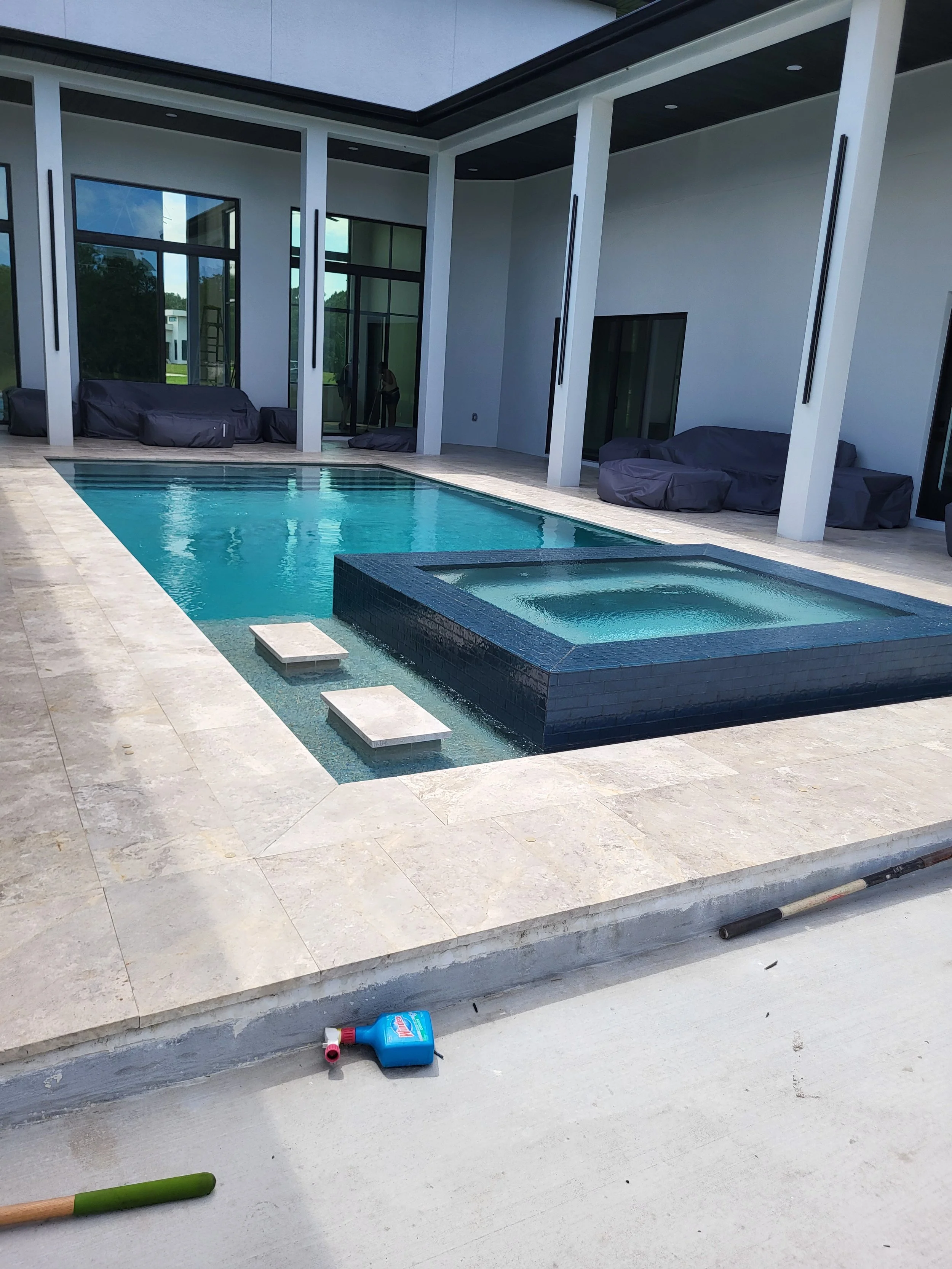Modern backyard with a swimming pool and hot tub, surrounded by a patio with covered outdoor furniture, overlooking a house with large glass doors and windows.