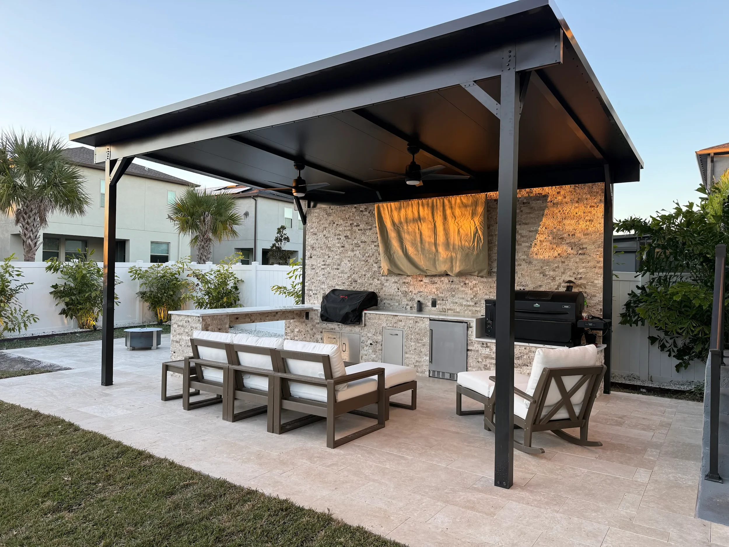 Outdoor patio with a covered roof, concrete flooring, seating area with cushioned chairs, a built-in grill, and decorative stone wall with a window covered by a tarp, surrounded by palm trees and greenery.
