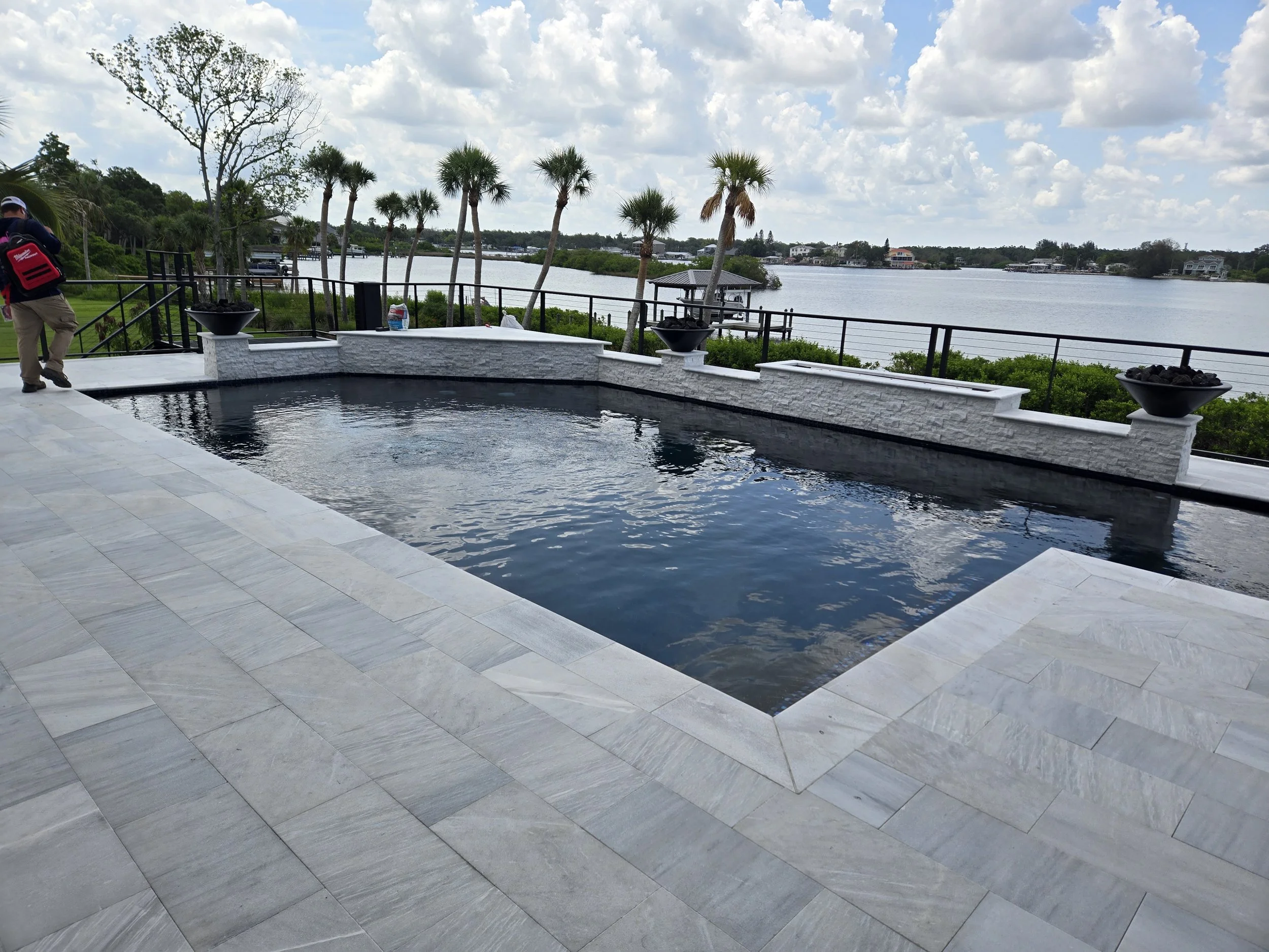 A backyard with a small rectangular swimming pool, a stone patio, palm trees, a river view, and a cloudy sky.