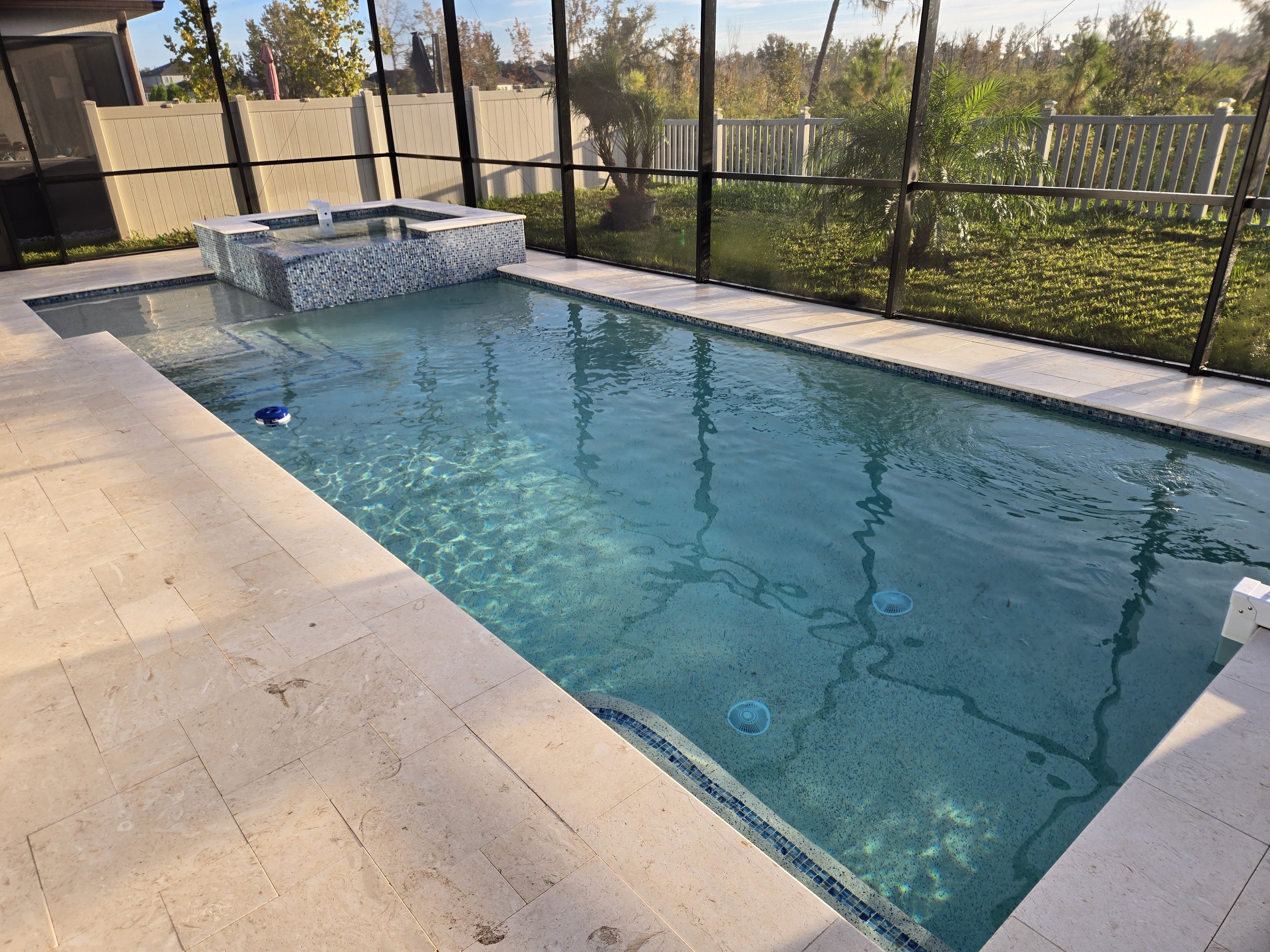 Backyard pool with a built-in hot tub and a screened enclosure, surrounded by a beige tile deck, with a green grassy yard, trees, and a white picket fence visible outside.