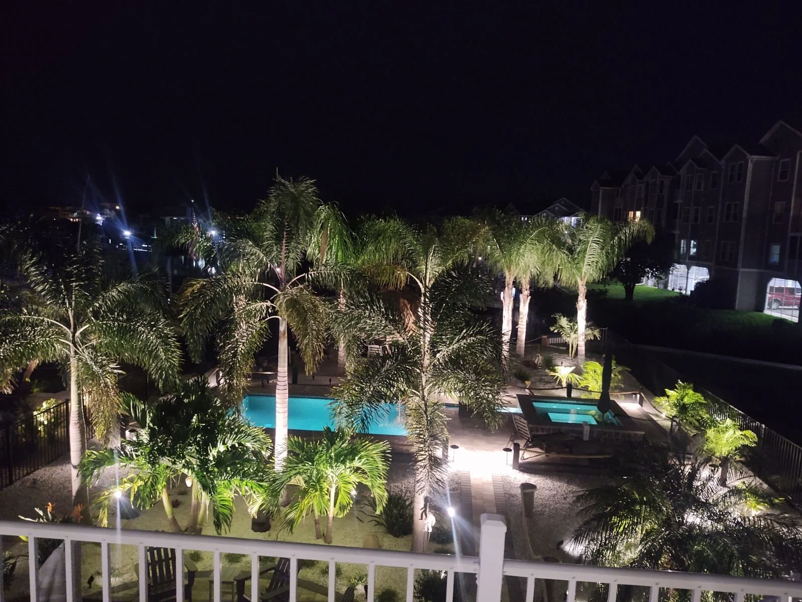 Nighttime view of a swimming pool area with tall palm trees, lit from below, surrounded by a patio, lounge chairs, and nearby apartment buildings.