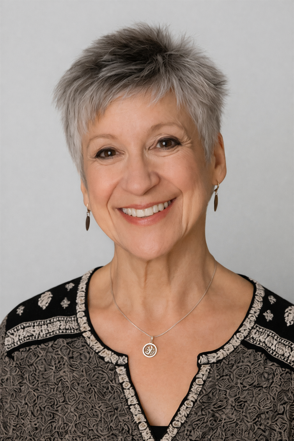 Portrait of a smiling older woman with short gray hair, wearing a black and silver patterned top and jewelry.