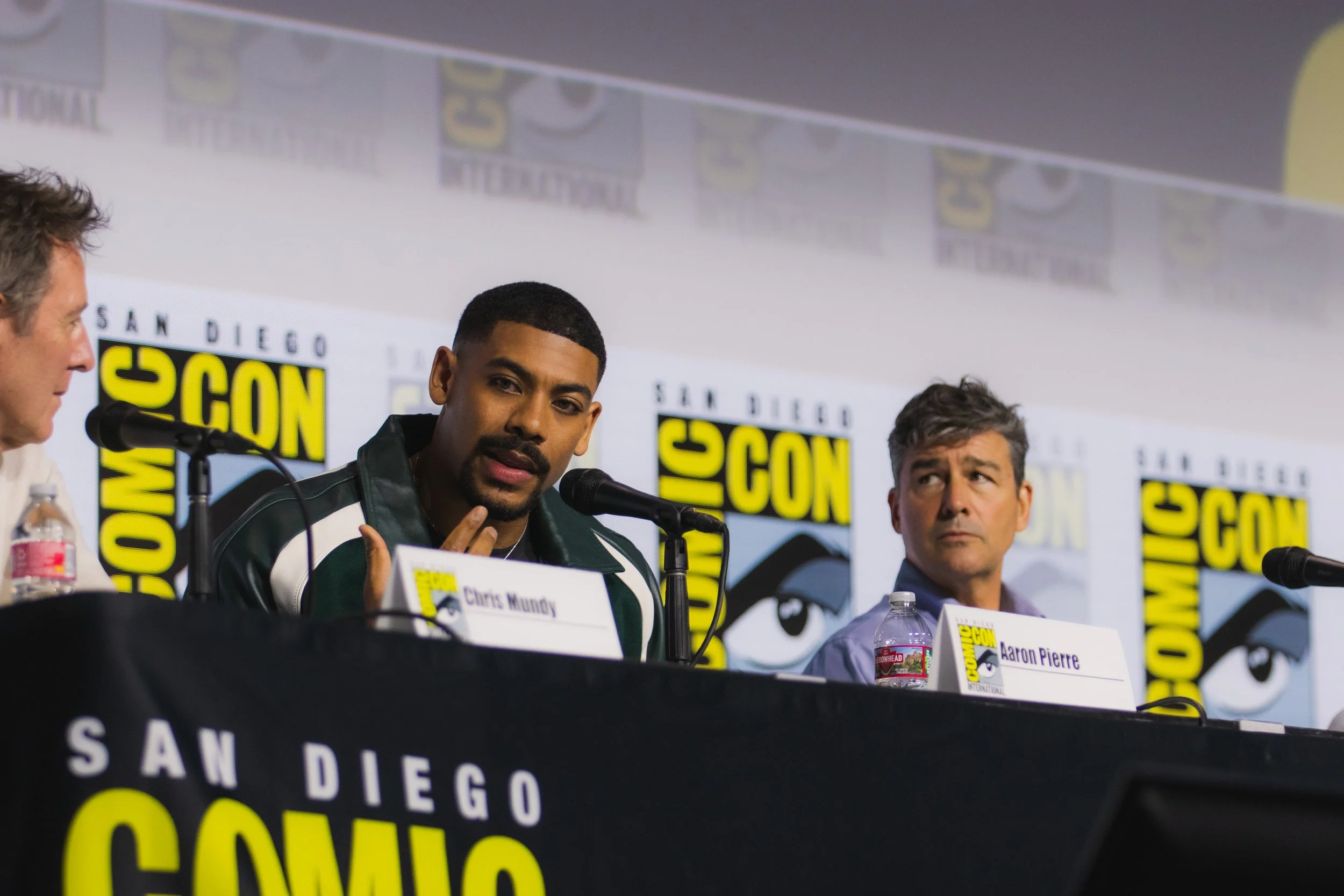 Aaron Pierre speaking into a microphone beside Kyle Chandler at a San Diego Comic-Con panel.