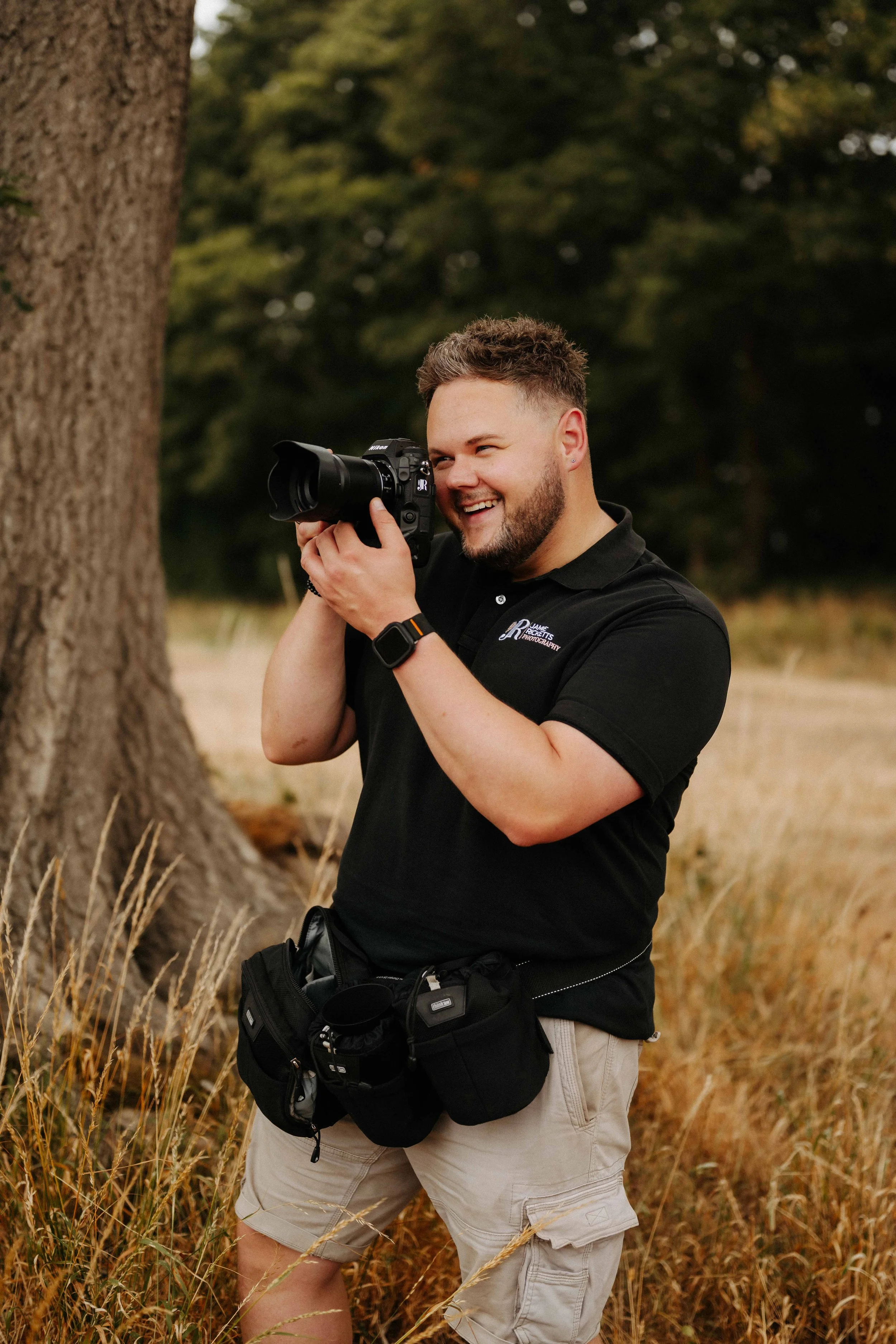 A smiling man taking a photograph outdoors next to a large tree, surrounded by tall dry grass and green trees in the background.