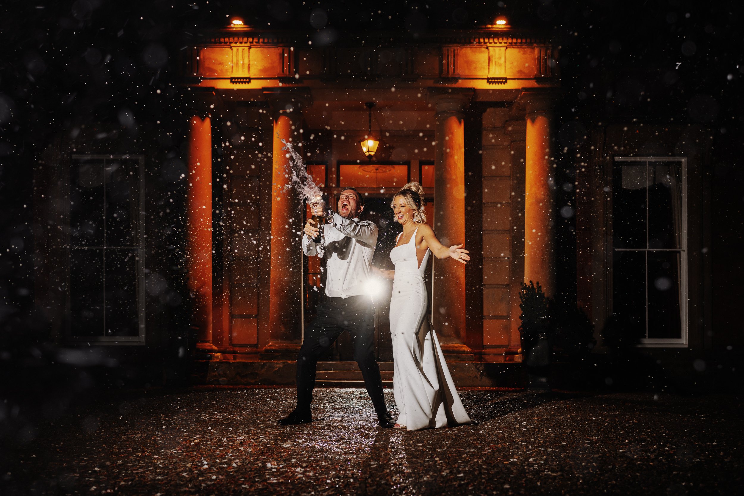 Bride and groom popping a bottle of champagne outside Netley Hall during their winter wedding celebration, spraying fizz and laughing together.