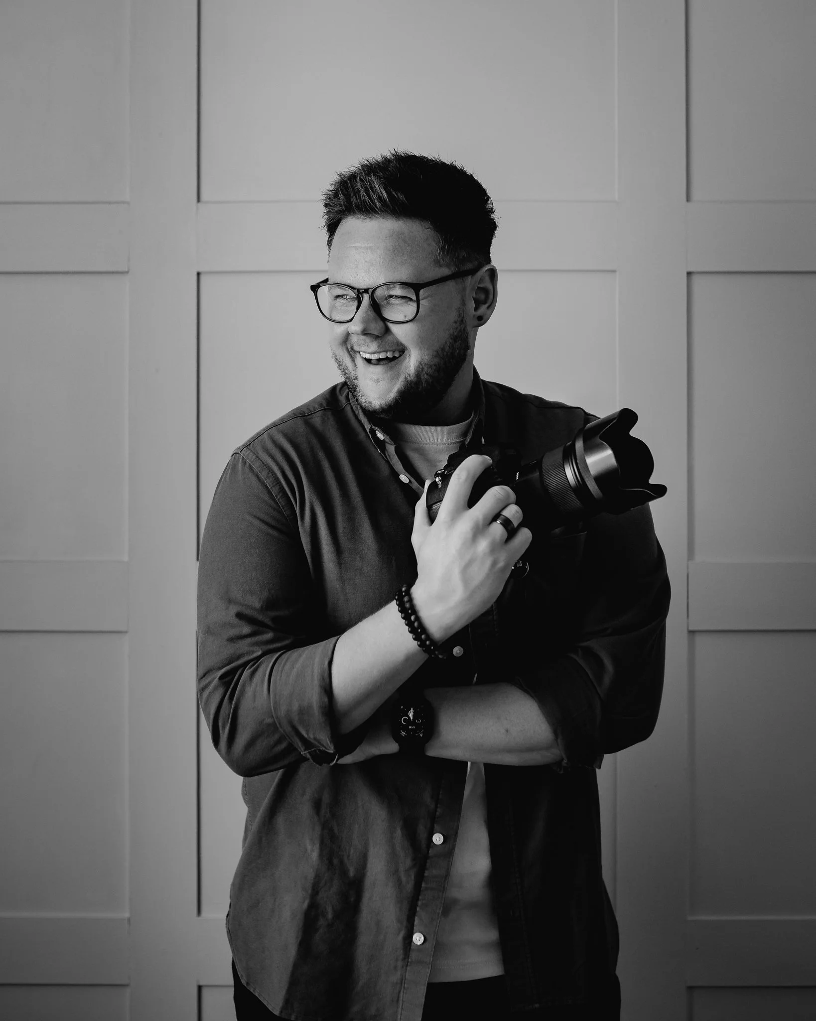 A smiling man with glasses and a beard holding a camera, standing in front of a paneled wall.