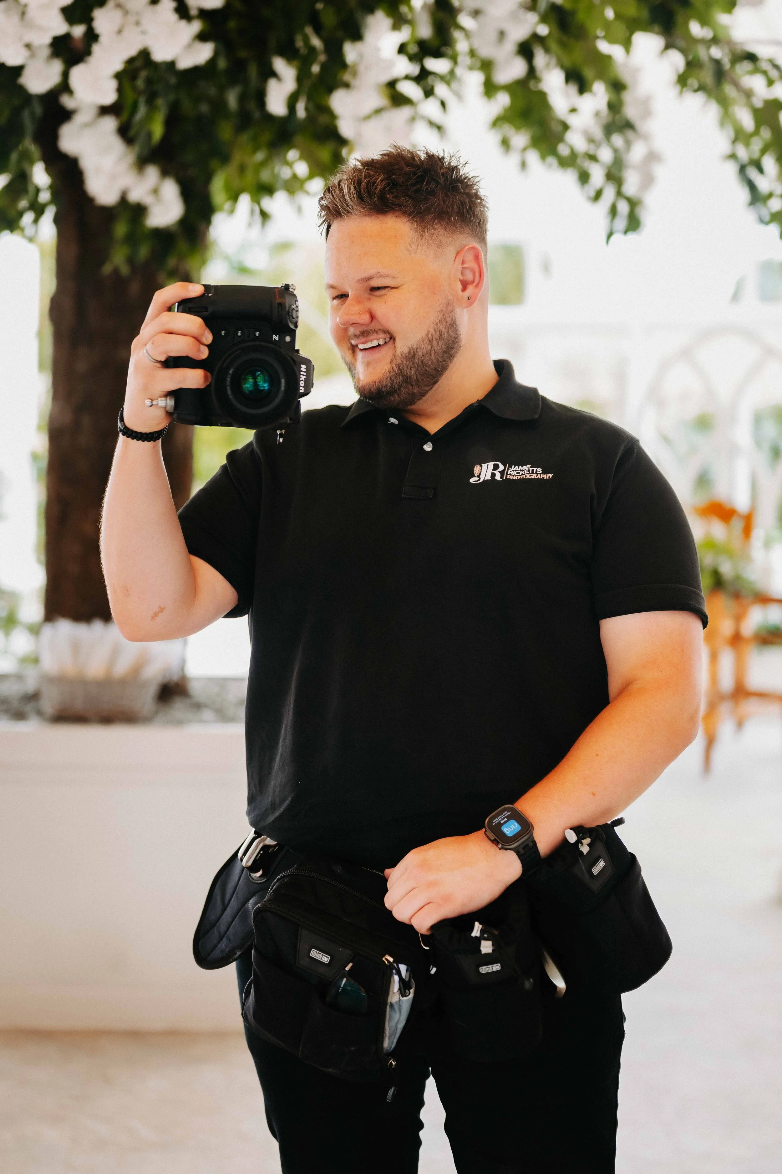 A smiling man in a black polo shirt with a logo, holding a camera. He has a beard and short, styled hair. The background features a blurred indoor setting with a large tree or plant and some decorative items.