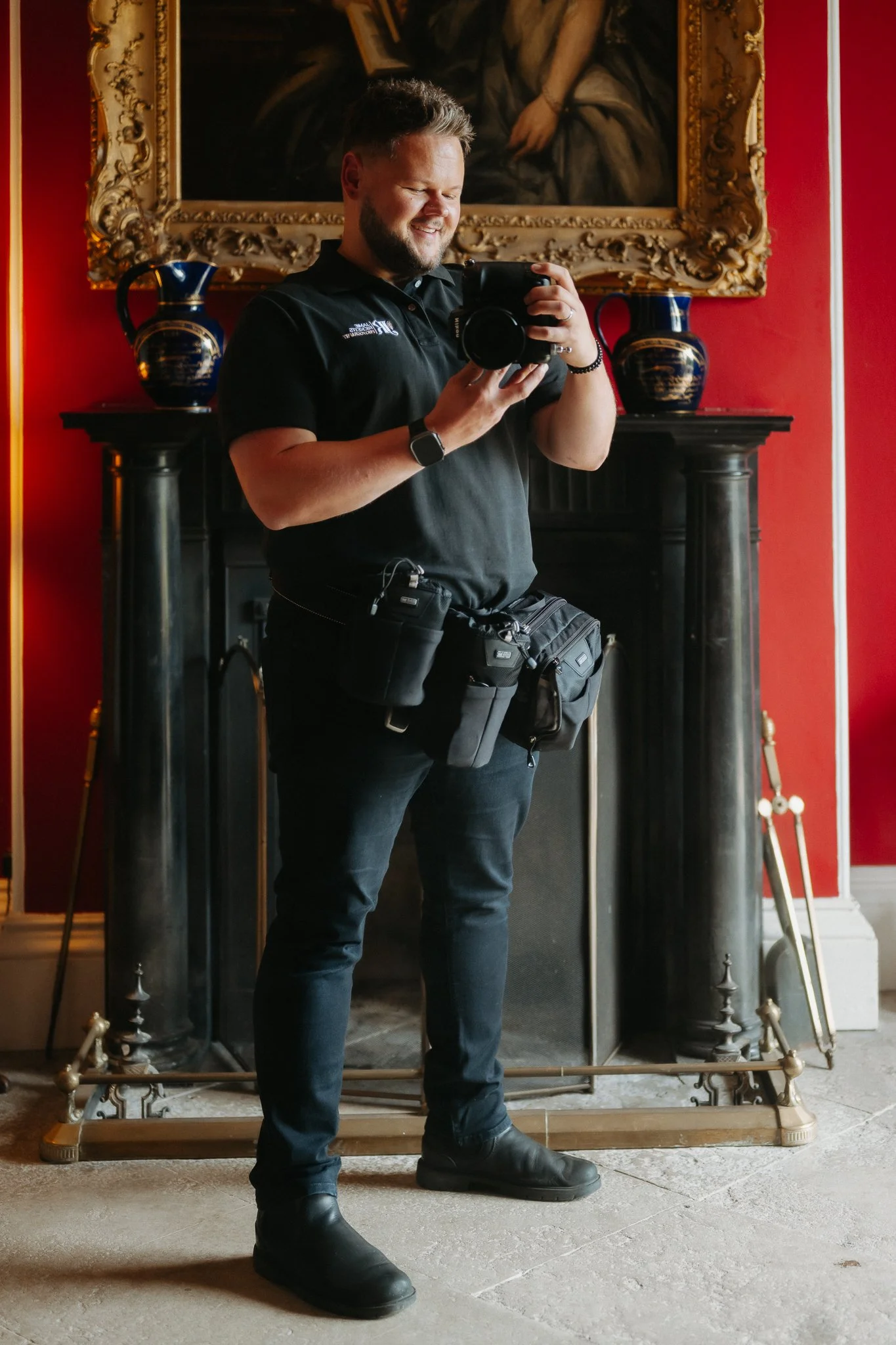 A man smiling while taking a selfie in a room with red walls, standing in front of a black fireplace with ornate gold and blue vases on either side and a large painting above.