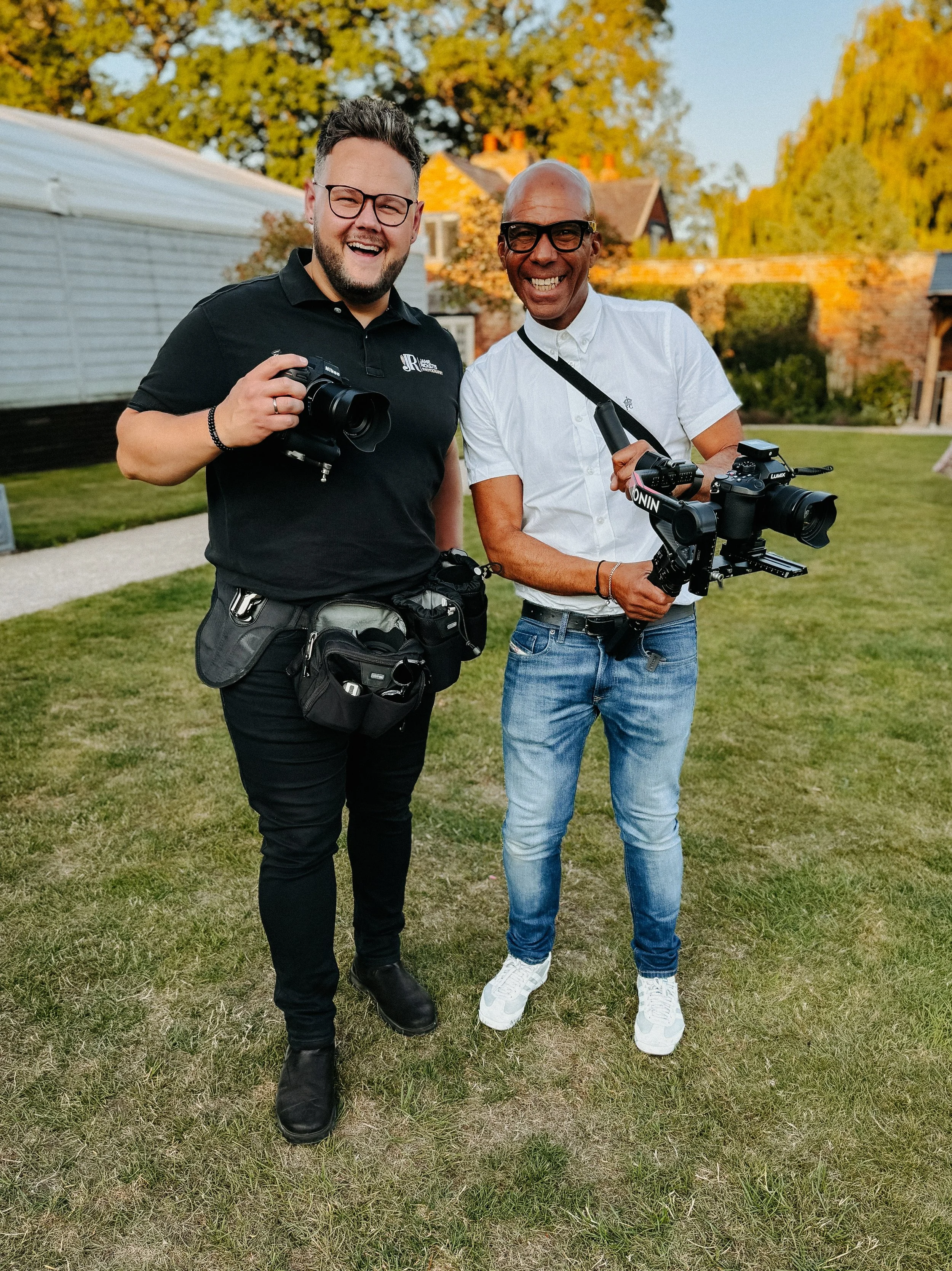 Two men standing outdoors on grass, smiling and holding cameras, with trees and houses in the background.