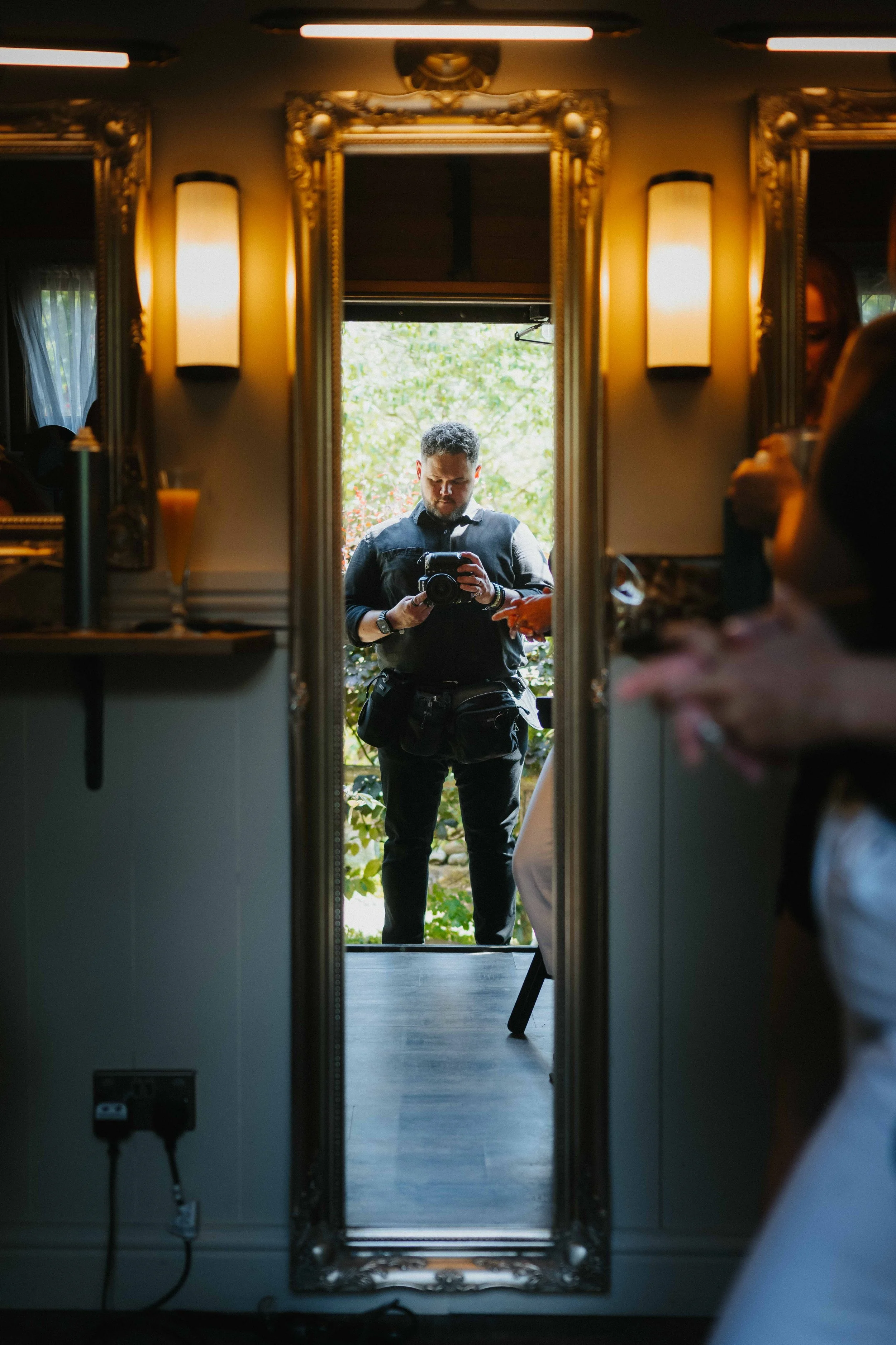 A man taking a photo of himself in a mirror outside, seen through an interior doorway with ornate gold trim, lit by warm wall lamps. The outdoor background shows trees and sunlight.
