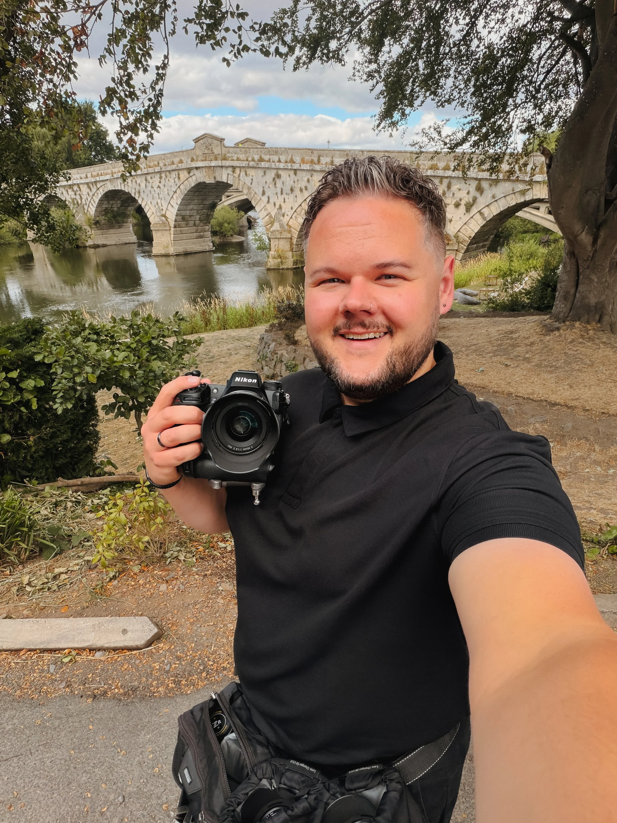 A man taking a selfie outdoors near a bridge over a river, holding a camera in his right hand, smiling, with trees and bushes around, and a cloudy sky overhead.
