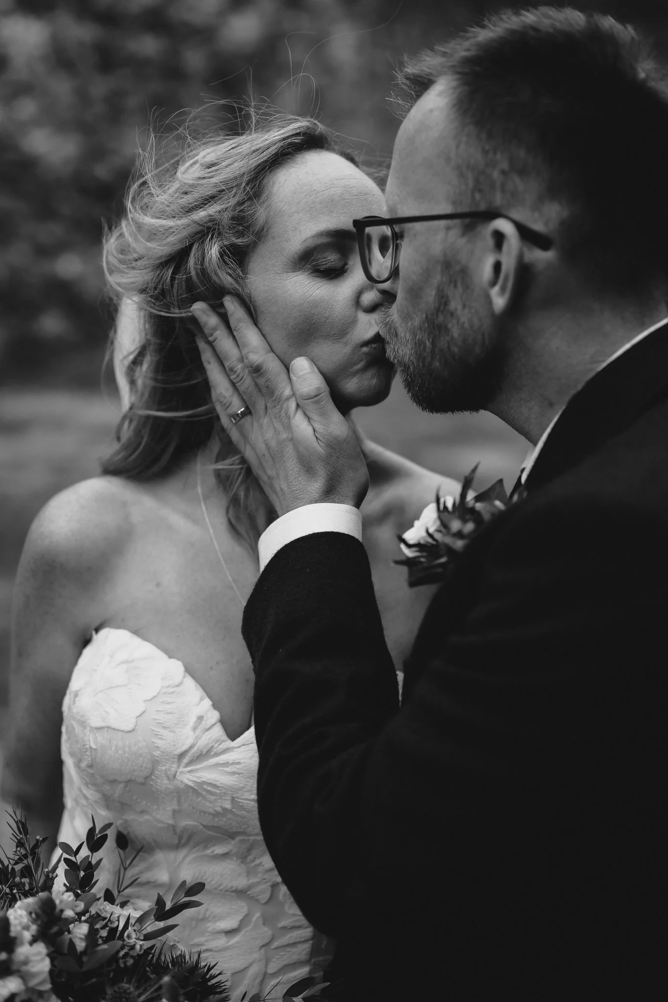 A black and white close-up of a couple sharing a kiss at their wedding. The woman has wavy hair and is wearing a strapless dress with floral details, and the man is wearing glasses and a tuxedo. The man's hand gently touches the woman's face, and there are flowers in the foreground.