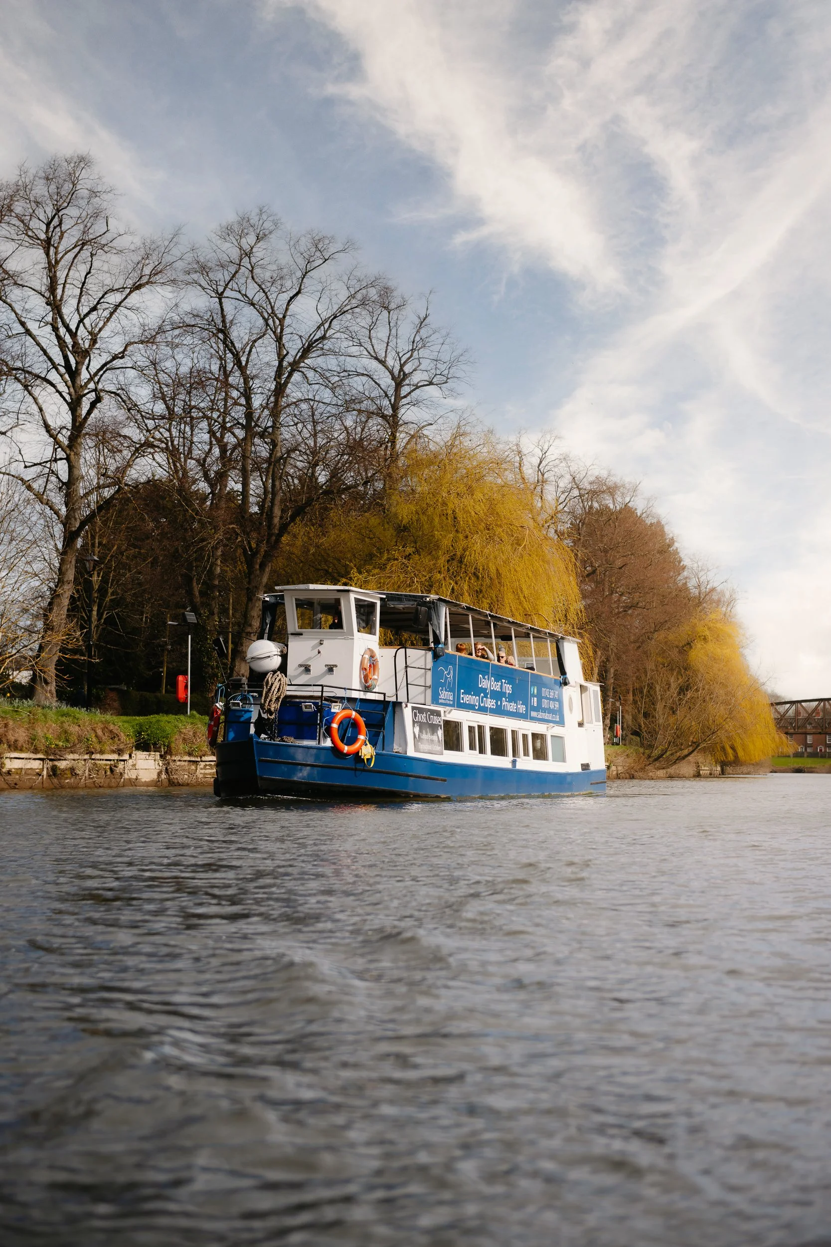 The Sabrina Boat carrying passengers along the River Severn in Shrewsbury, with the open green space of Shrewsbury Quarry visible in the background in Shropshire.