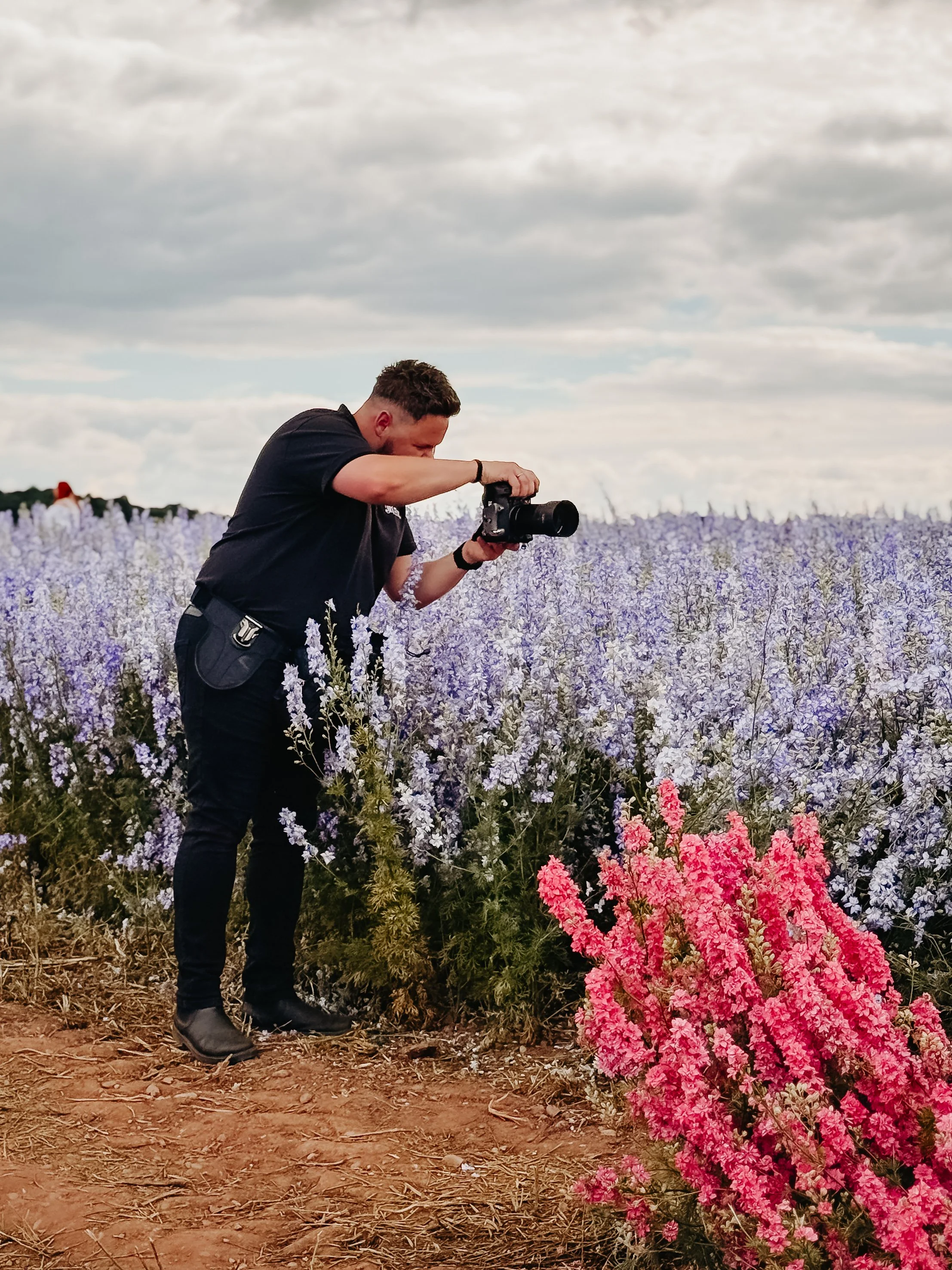 A man in black clothing and boots is taking a photograph in a field of purple and pink flowers under a cloudy sky.