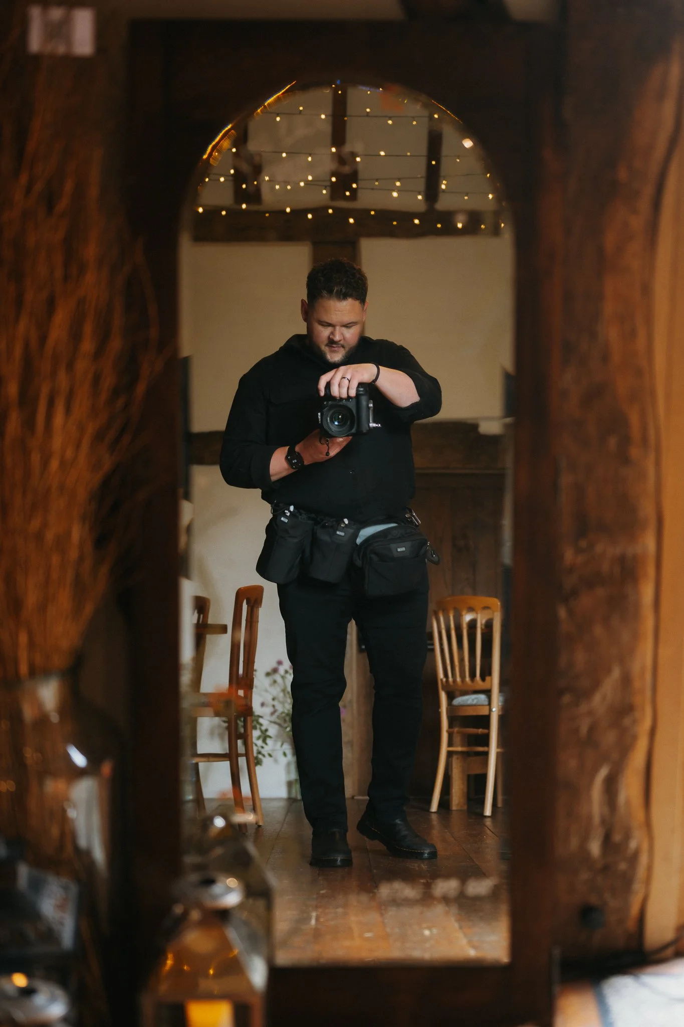 A man dressed in black taking a photo of himself in a mirror framed by wooden furniture, with string lights illuminating the background.