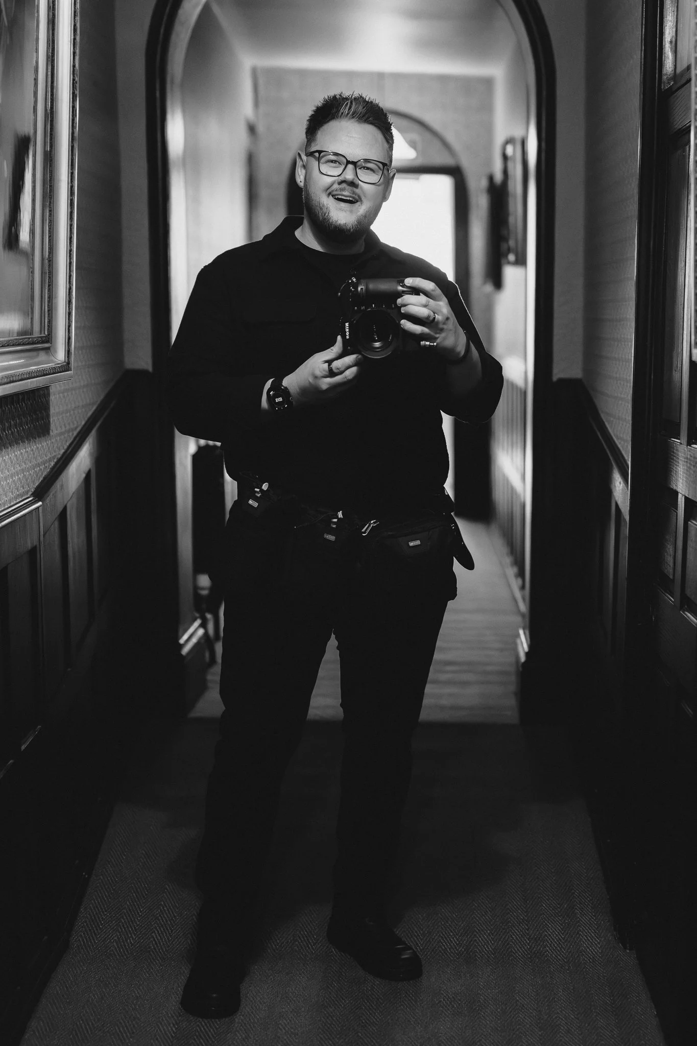 A young man with glasses and a beard takes a selfie in a mirror in a hallway, holding a camera with a smile.