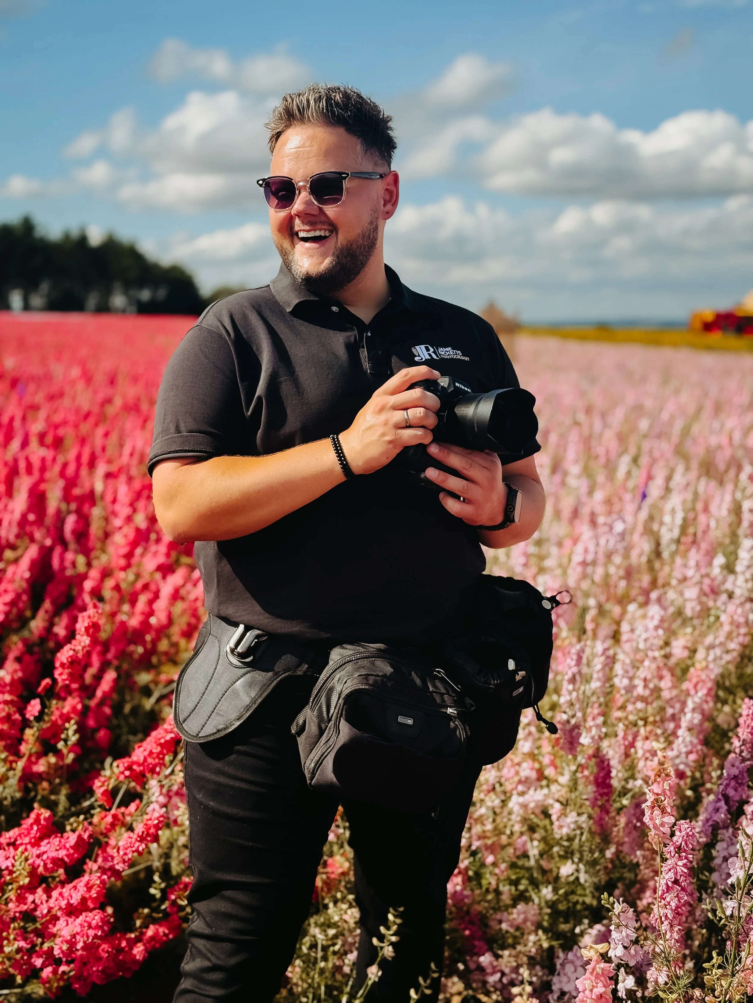 A man with sunglasses, a beard, and a black polo shirt stands outdoors holding a camera, smiling, in a field of colorful flowers under a partly cloudy sky.