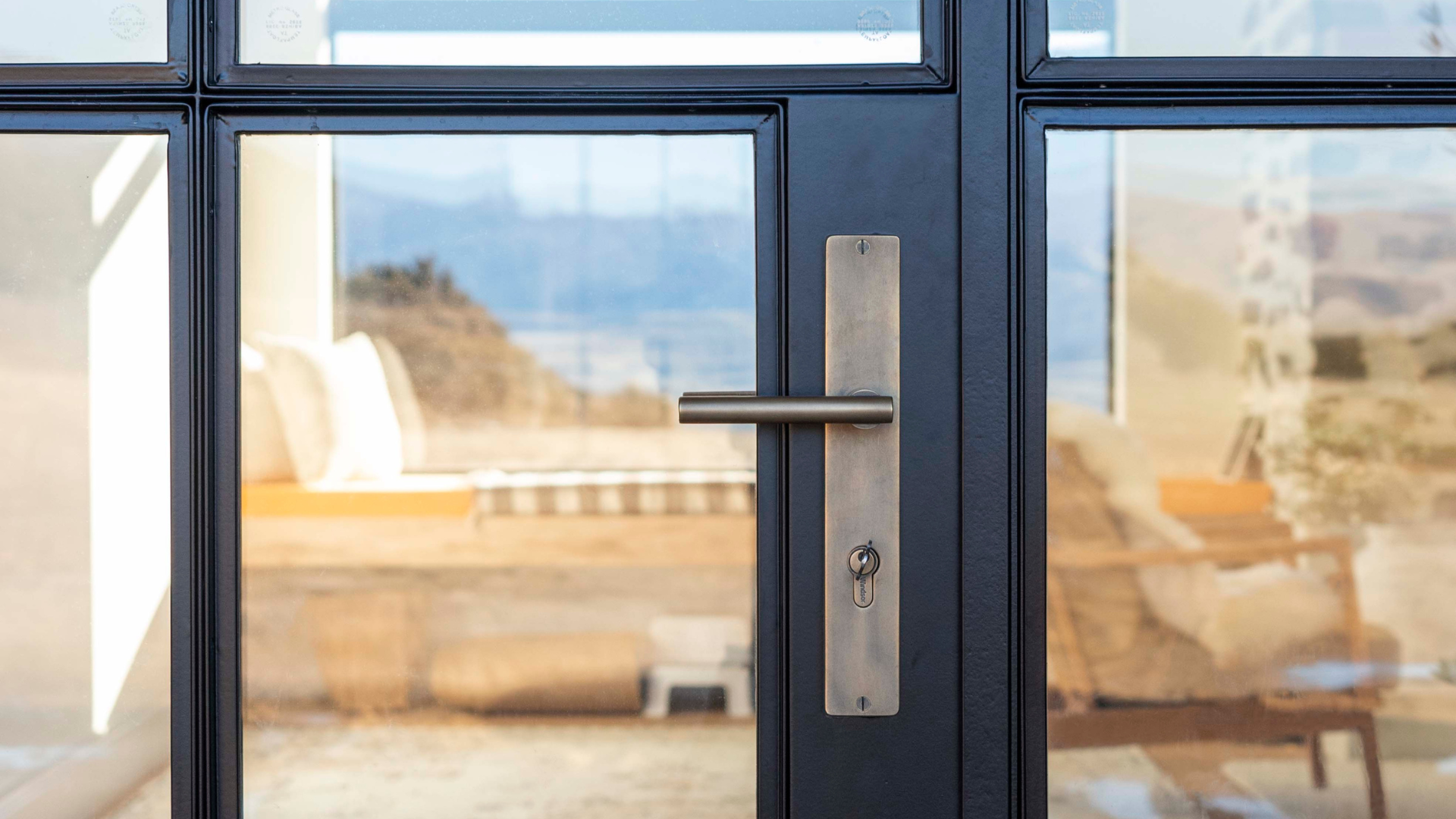 Close-up view of a modern glass door with a metal handle created by Southland company Windsor Architectural Hardware , lock, and keyhole, through which a scenic outdoor landscape with mountains and blue sky is visible.
