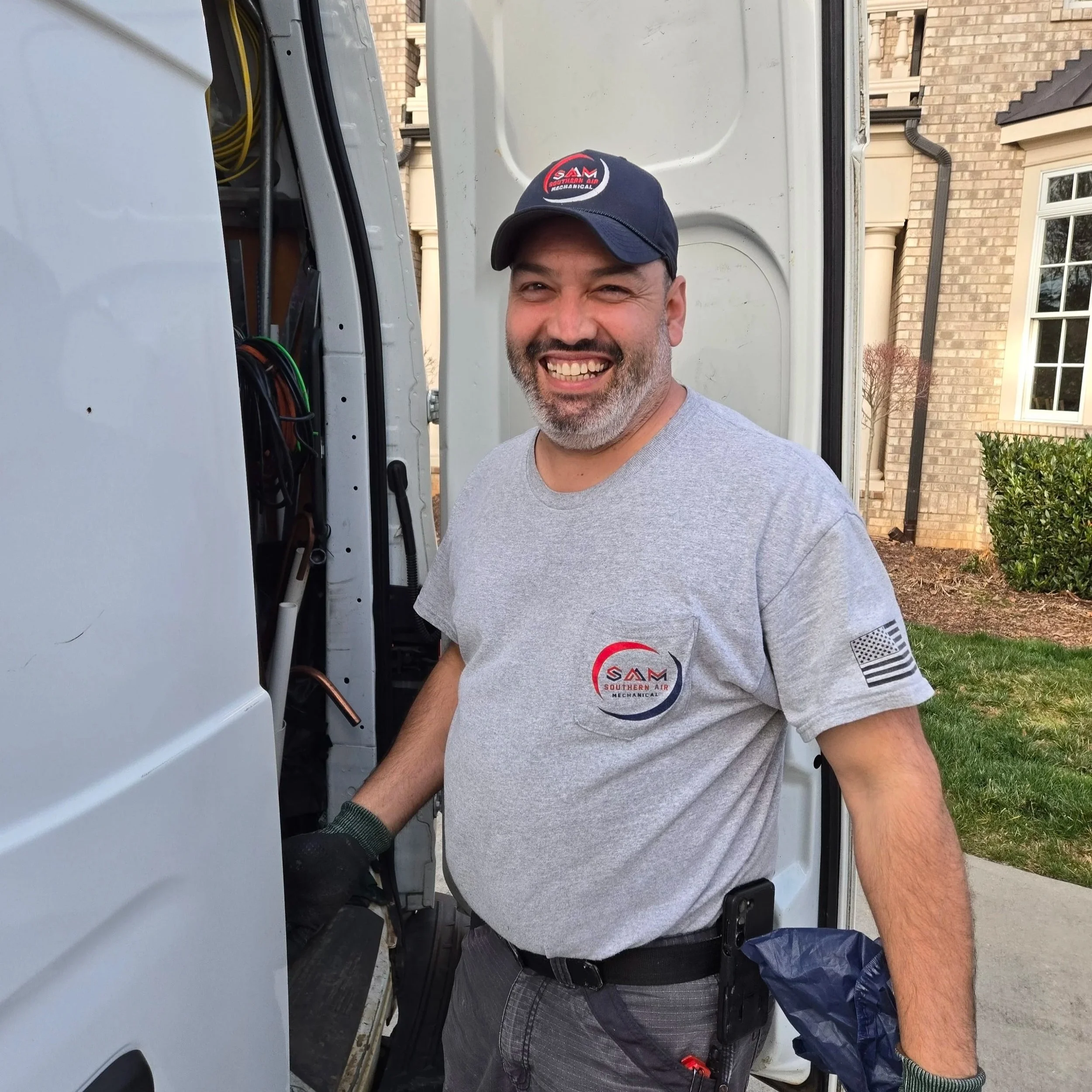 A man with a beard wearing a gray T-shirt, black gloves, and a navy cap with a red and white logo, working on a mechanical or electrical task outside near a beige brick house and a white utility truck.