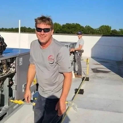 Two men working on a rooftop with HVAC or electrical equipment, one smiling at the camera and wearing sunglasses.