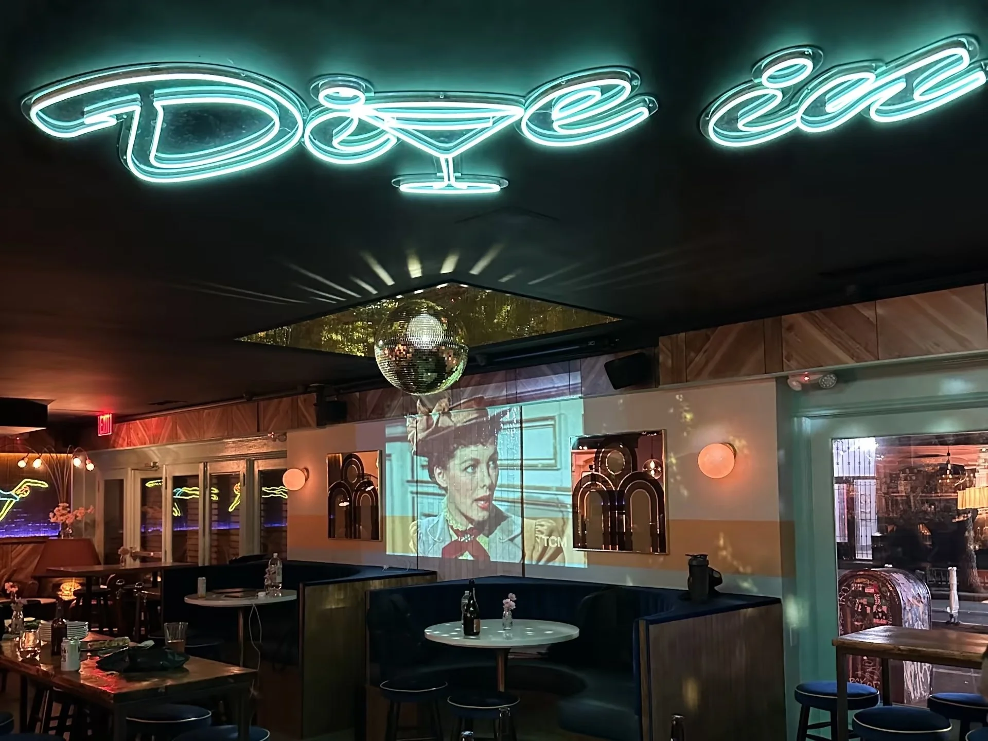 Interior of a bar or restaurant with a neon sign reading 'Dance' on the ceiling, a disco ball, and a large screen playing a vintage film with a woman on it.