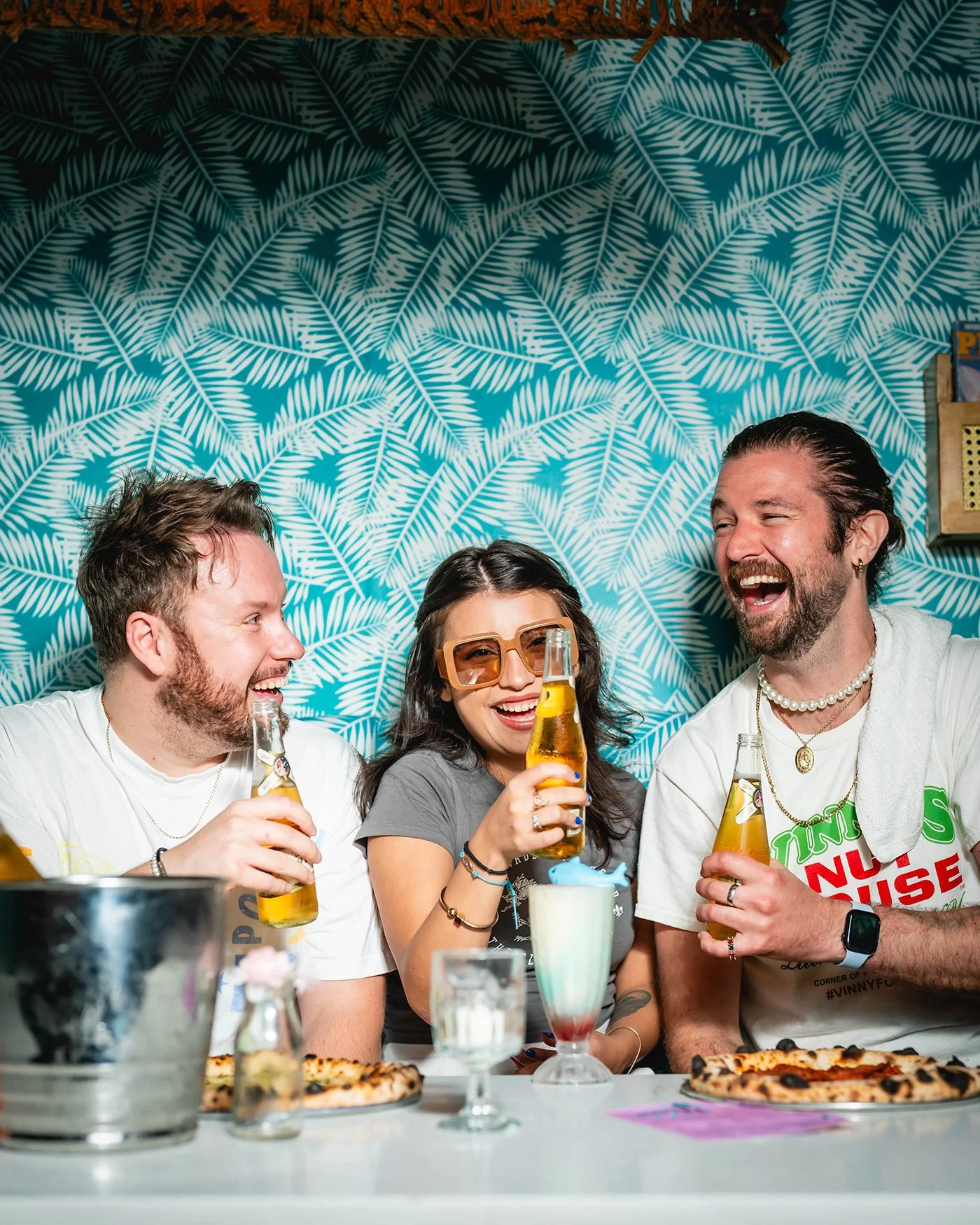 Three friends enjoying drinks and pizza at a table, laughing and smiling together.