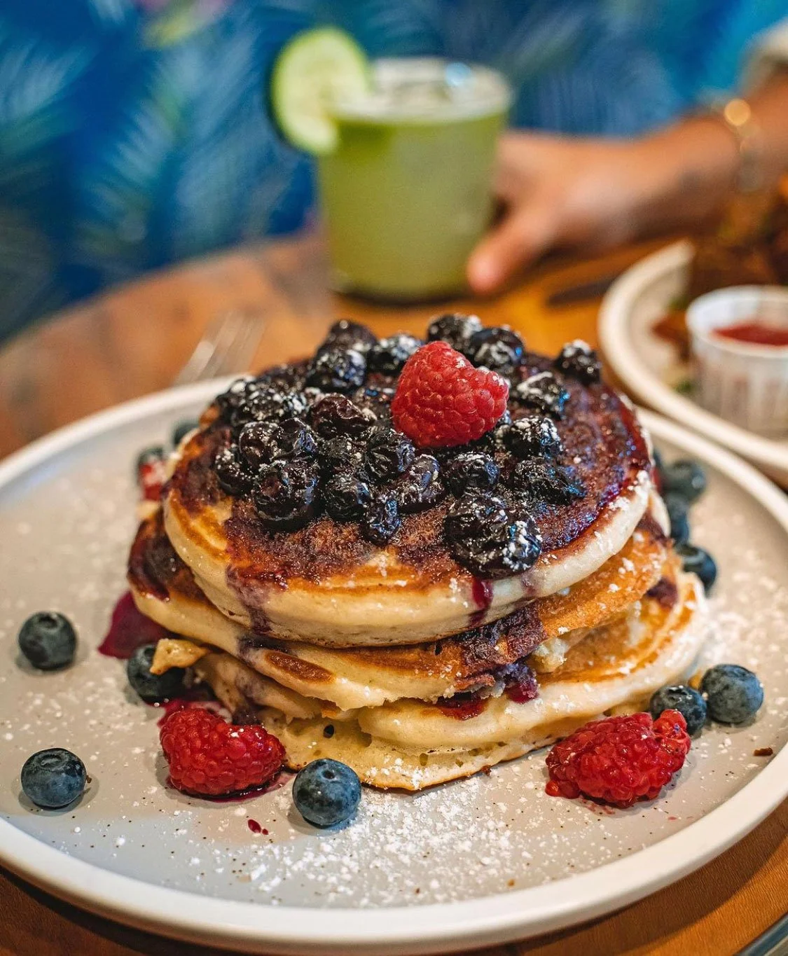 Stack of pancakes topped with blueberries, raspberries, and syrup, dusted with powdered sugar, on a white plate with a green drink in the background.