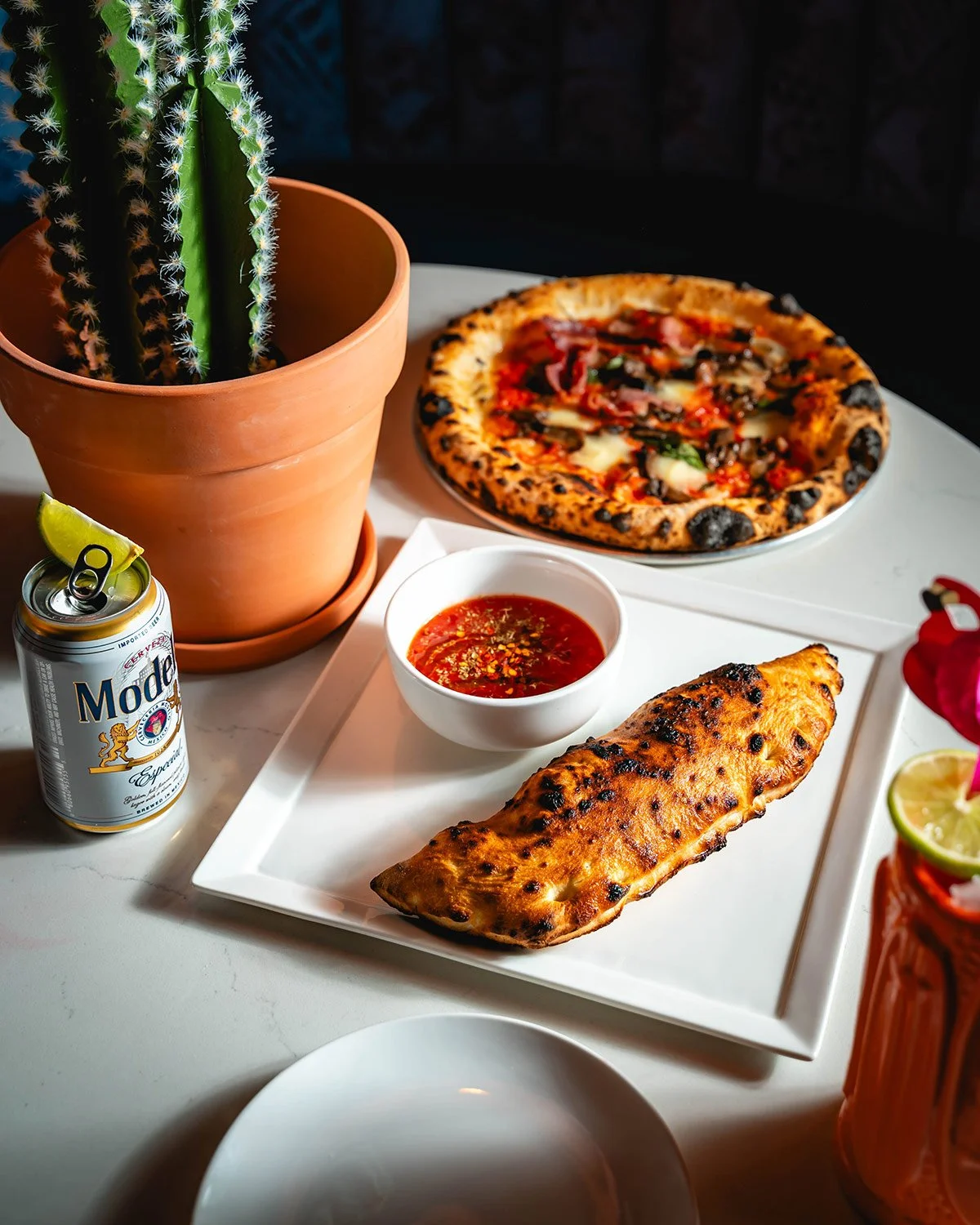 A table with a potted cactus, a pizza, a calzone, a bowl of marinara sauce, a can of Modelo beer, and a colorful drink with lime and a flower.
