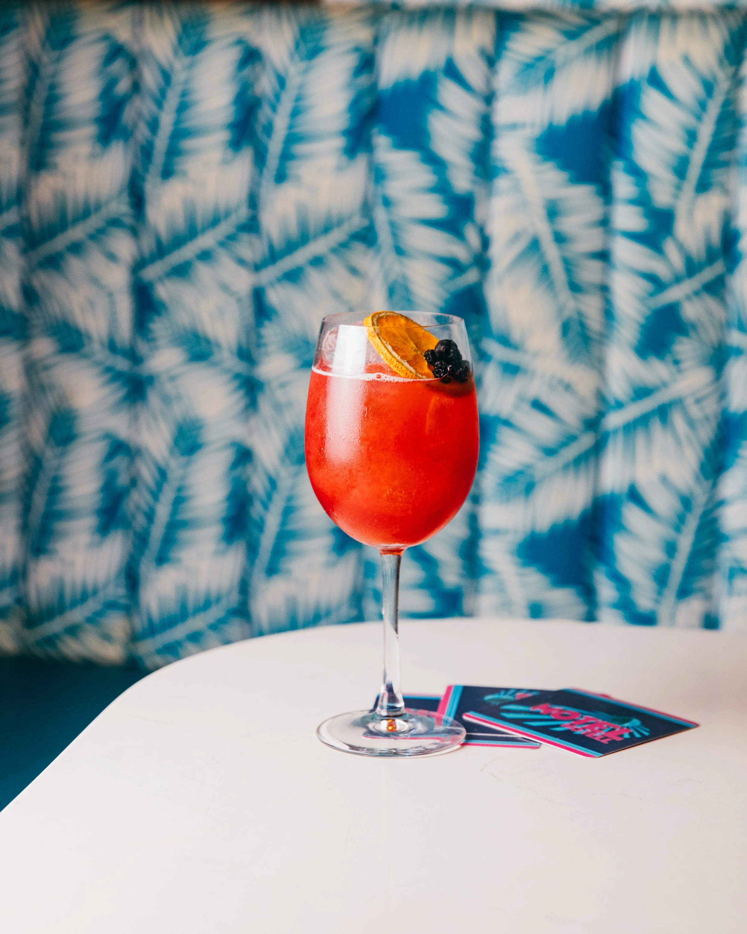A red cocktail in a wine glass garnished with a dried orange slice and blackberries, set on a white table with colorful cards nearby, against a background of blue and white patterned fabric.