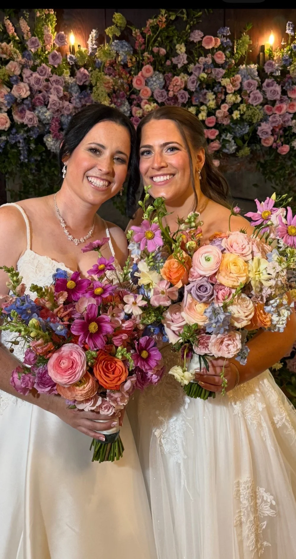 Two brides holding colorful bouquets in front of a flower wall