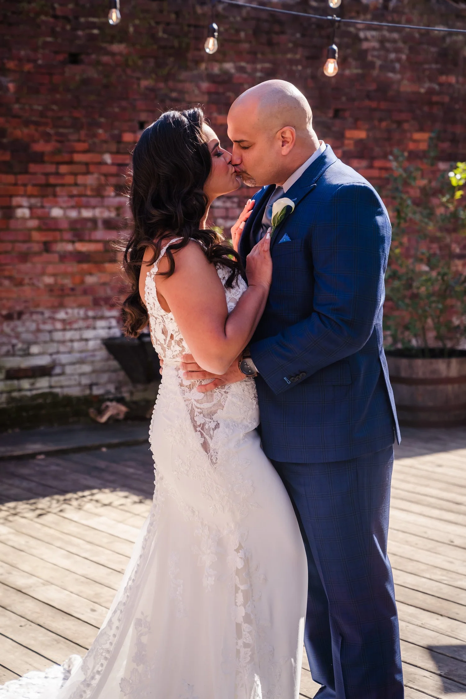 Bride and groom embracing under string lights by a brick wall