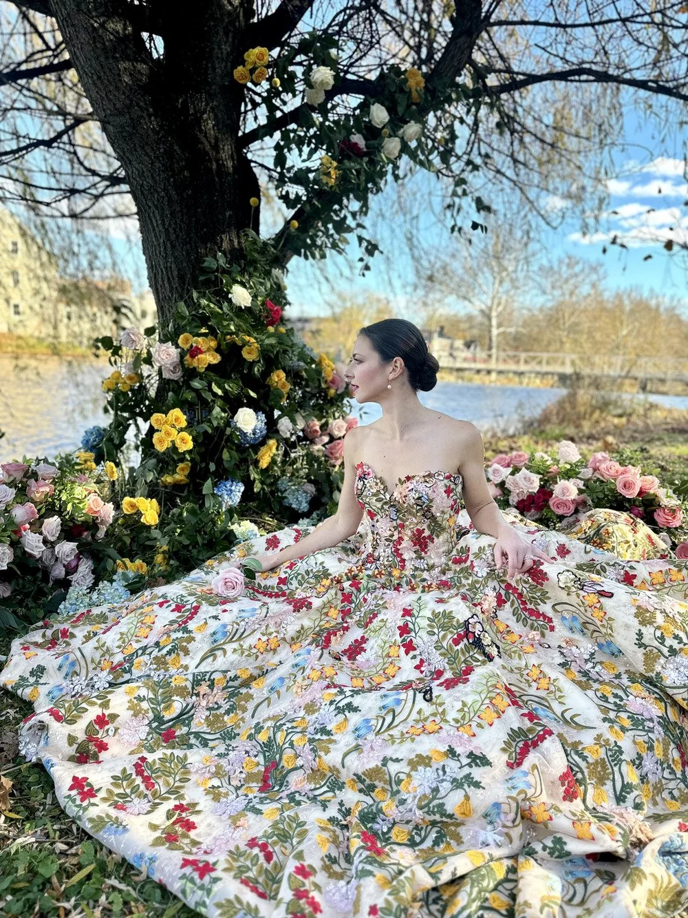 Woman in a floral gown seated under a flower-covered tree by the river