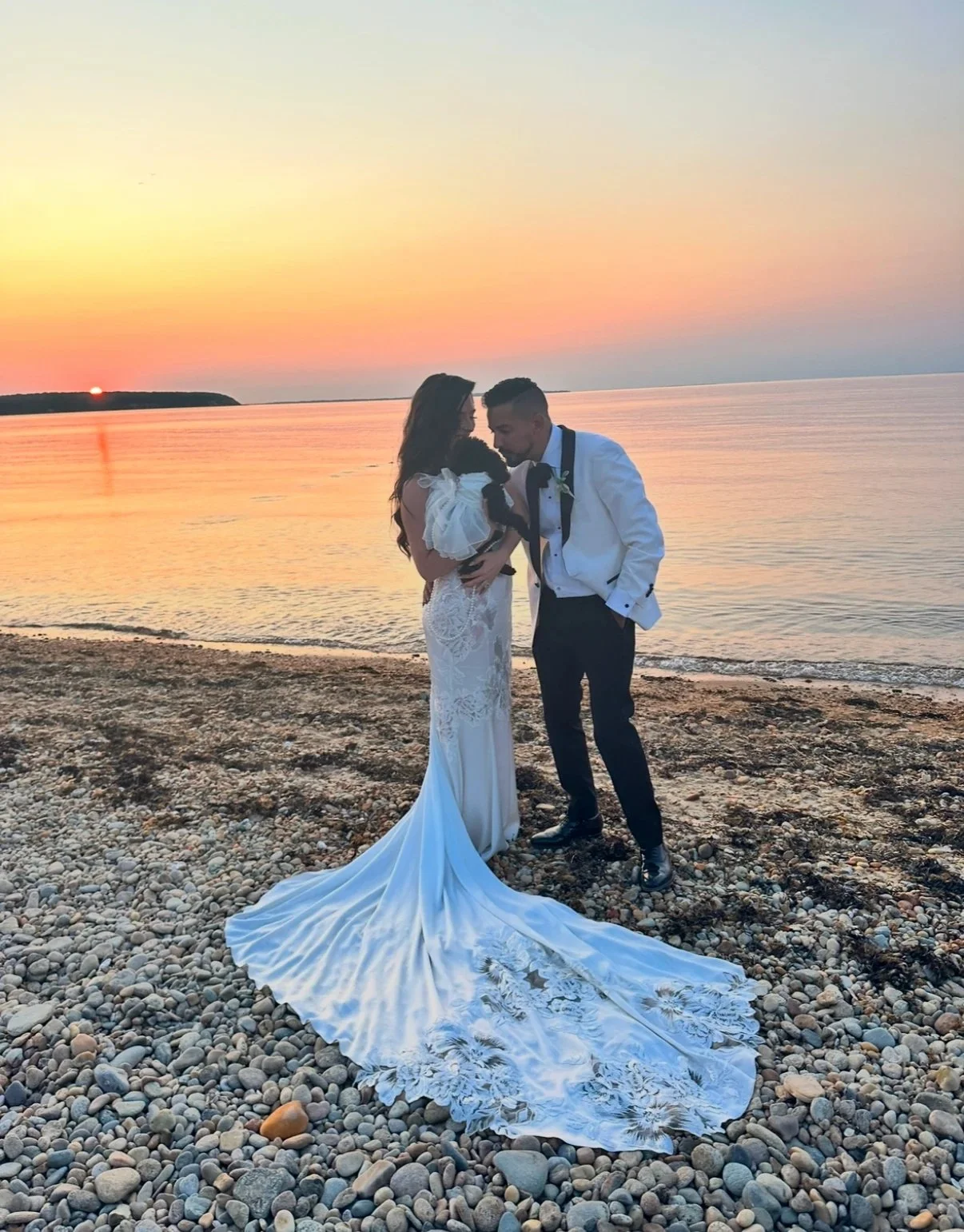 Bride and groom holding their dog on a pebble beach at sunset