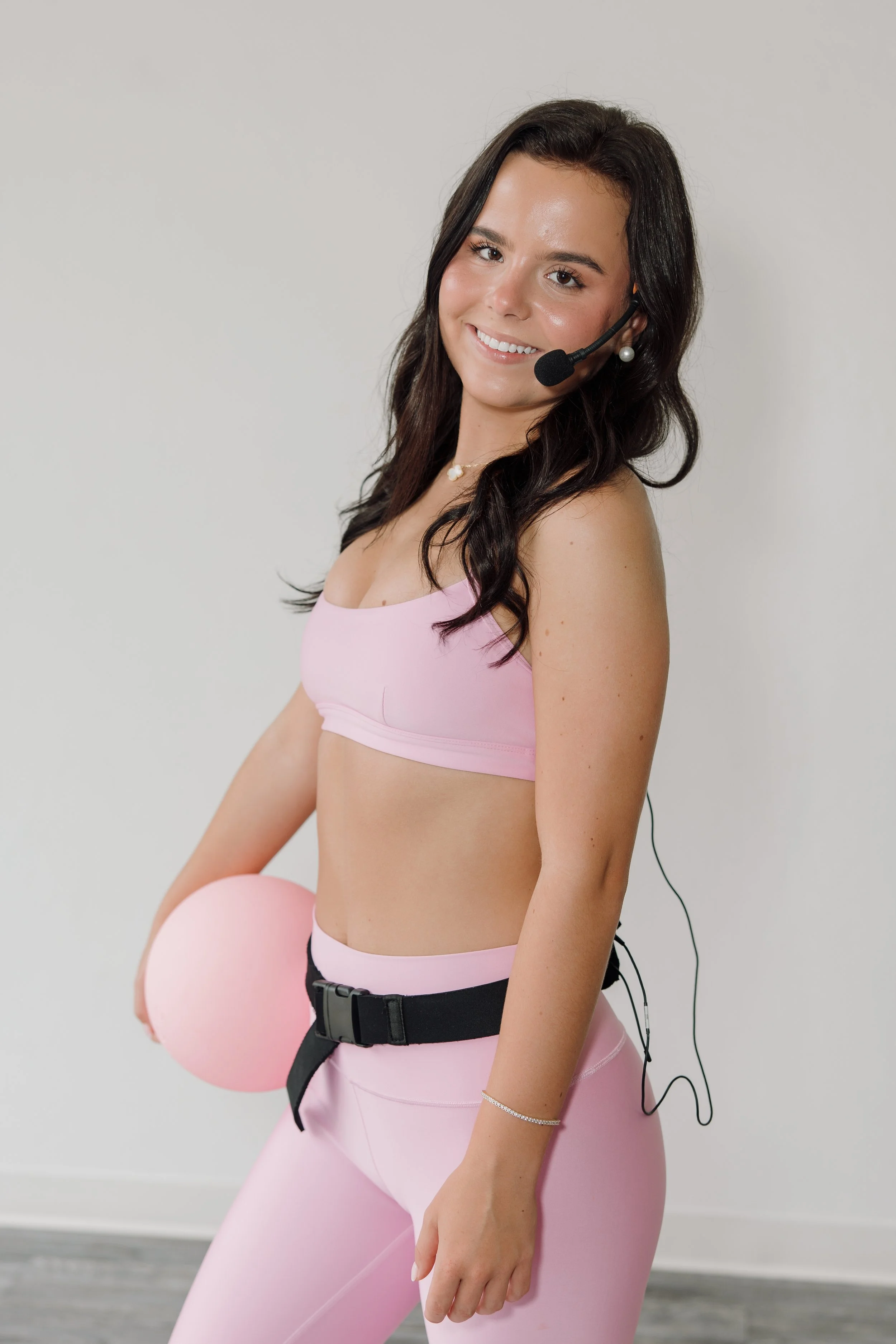 Young woman with dark hair, wearing a pink sports bra and leggings, smiling, holding a pink balloon, and wearing a headset with a microphone.