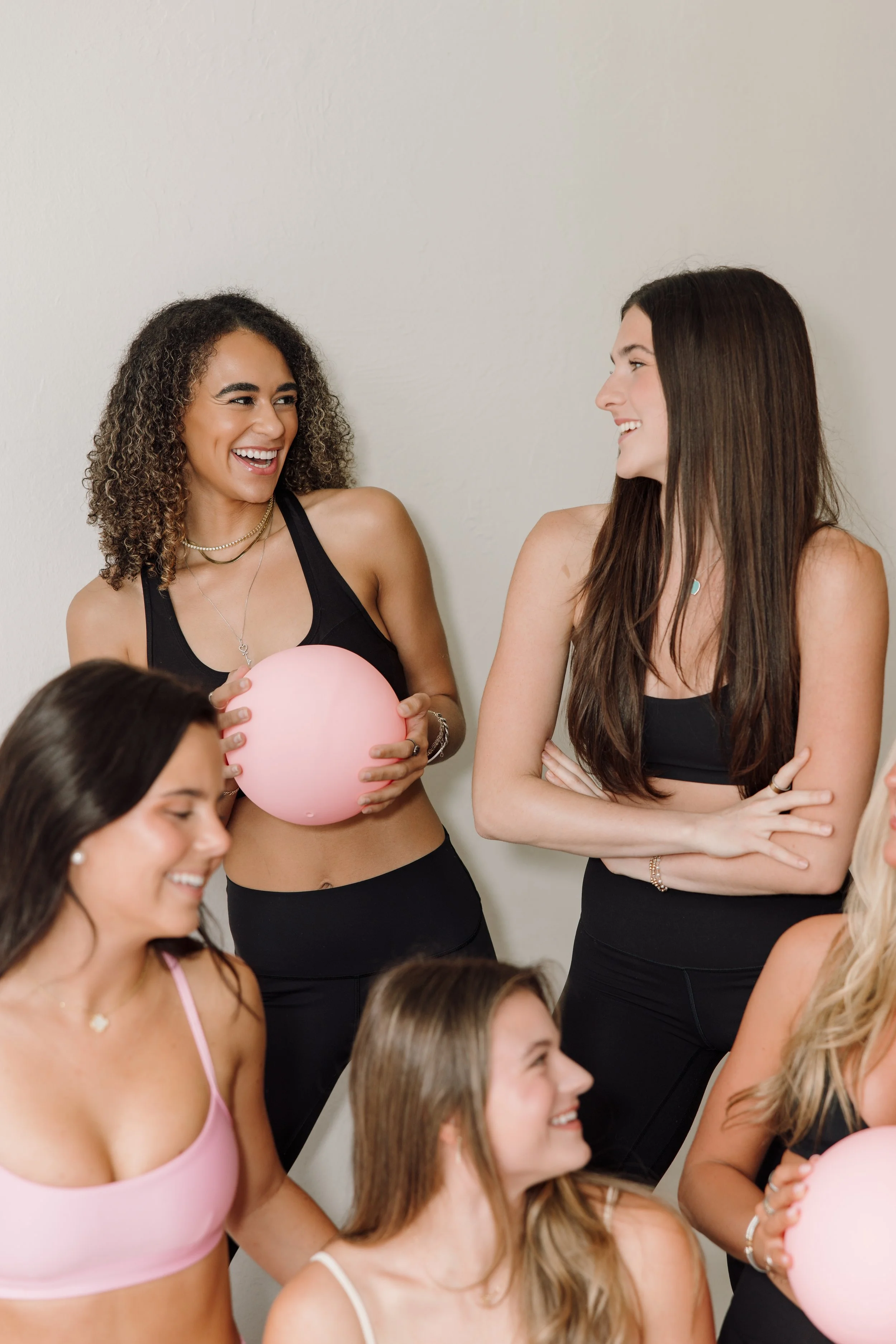 Group of women at a party smiling and talking, some holding pink balloons, dressed in casual tank tops and dresses.