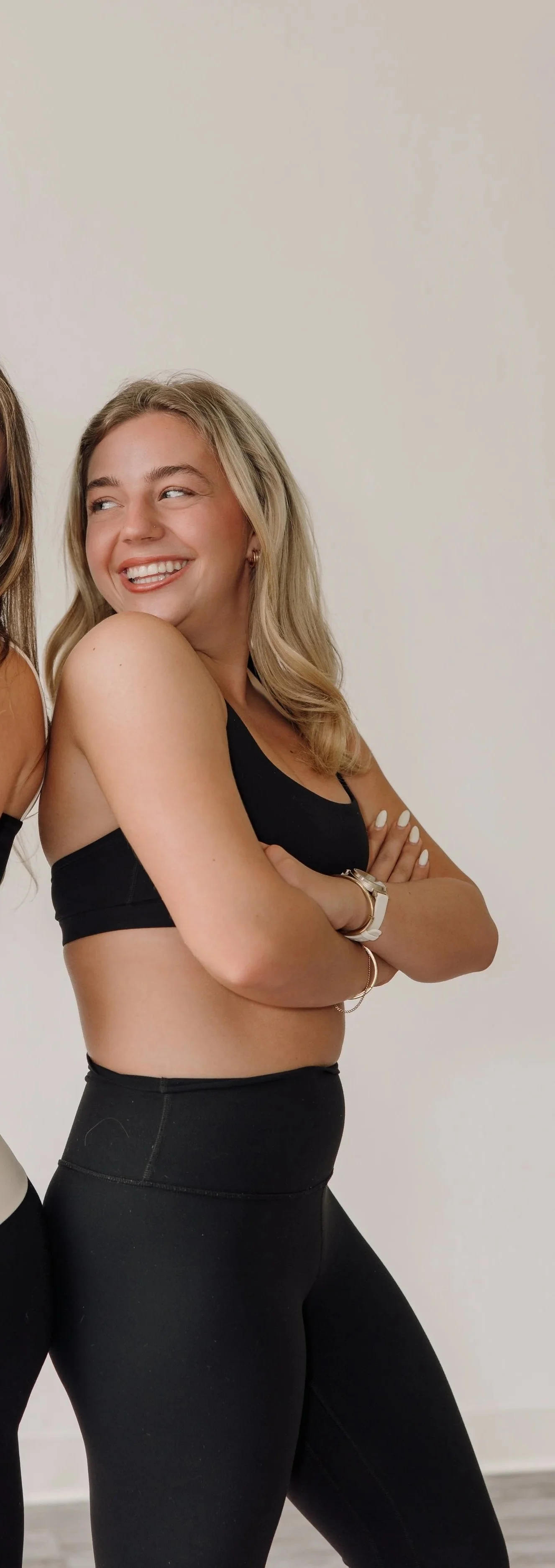 A woman with blonde hair smiling with crossed arms, wearing black workout clothes, standing against a plain white wall.