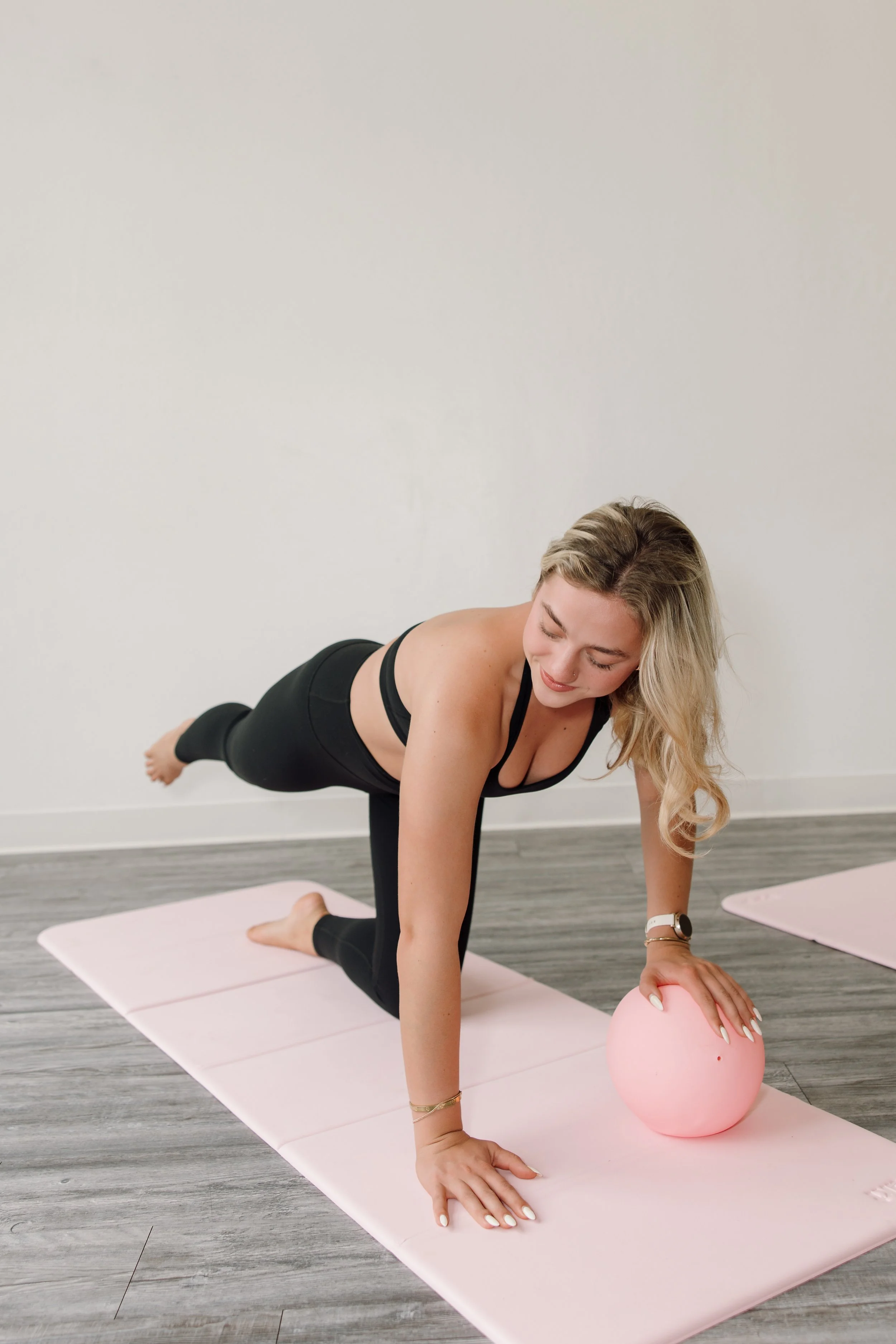 A woman practicing yoga on a pink mat, reaching for a pink exercise ball, in a room with wood flooring and plain white walls.