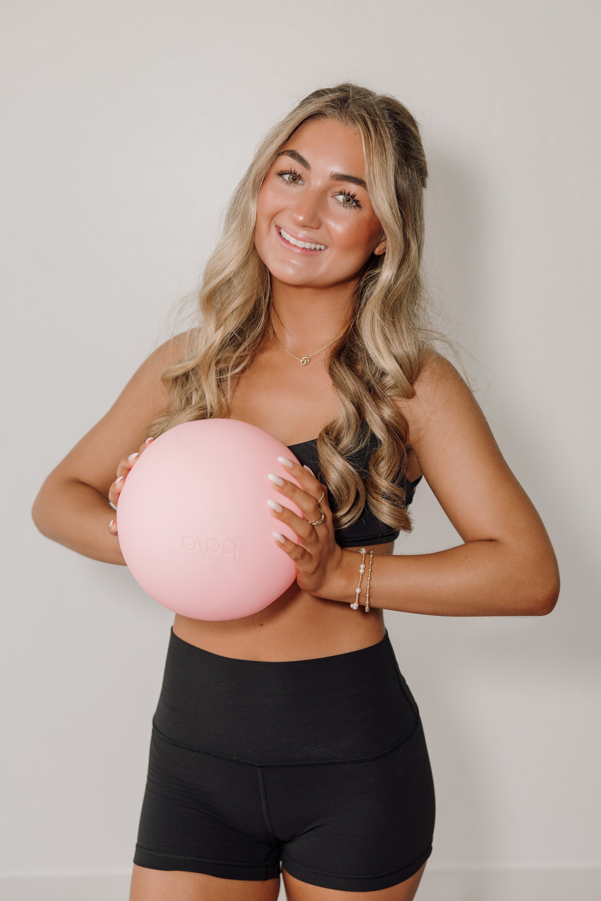 A woman with long blonde hair in loose waves, smiling, holding a light pink exercise ball, wearing a black sports bra and high-waisted black shorts, standing against a plain light-colored wall.