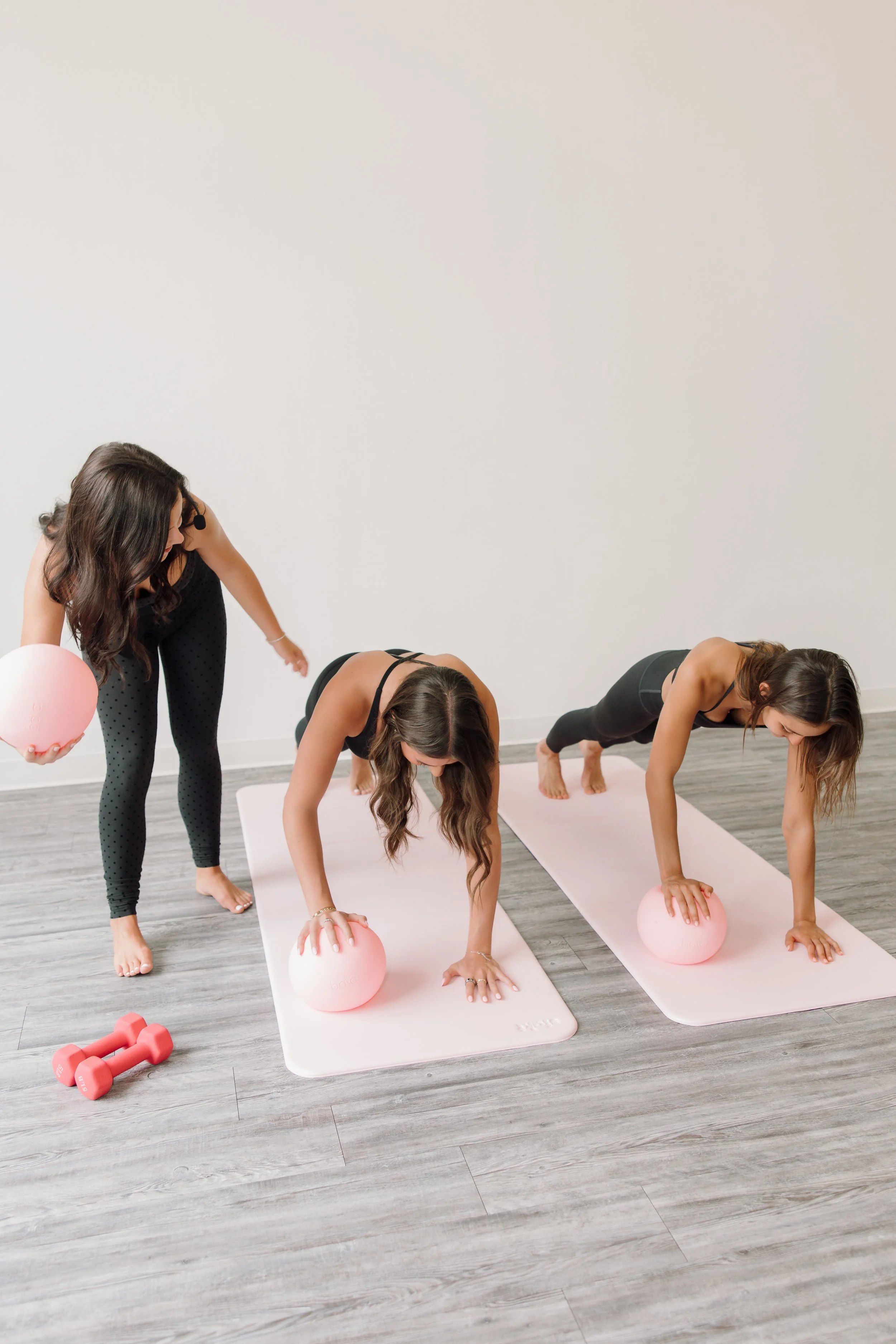 Three women doing a yoga workout in a fitness studio, on pink mats with pink exercise balls, while a trainer holds a pink ball and guides them.