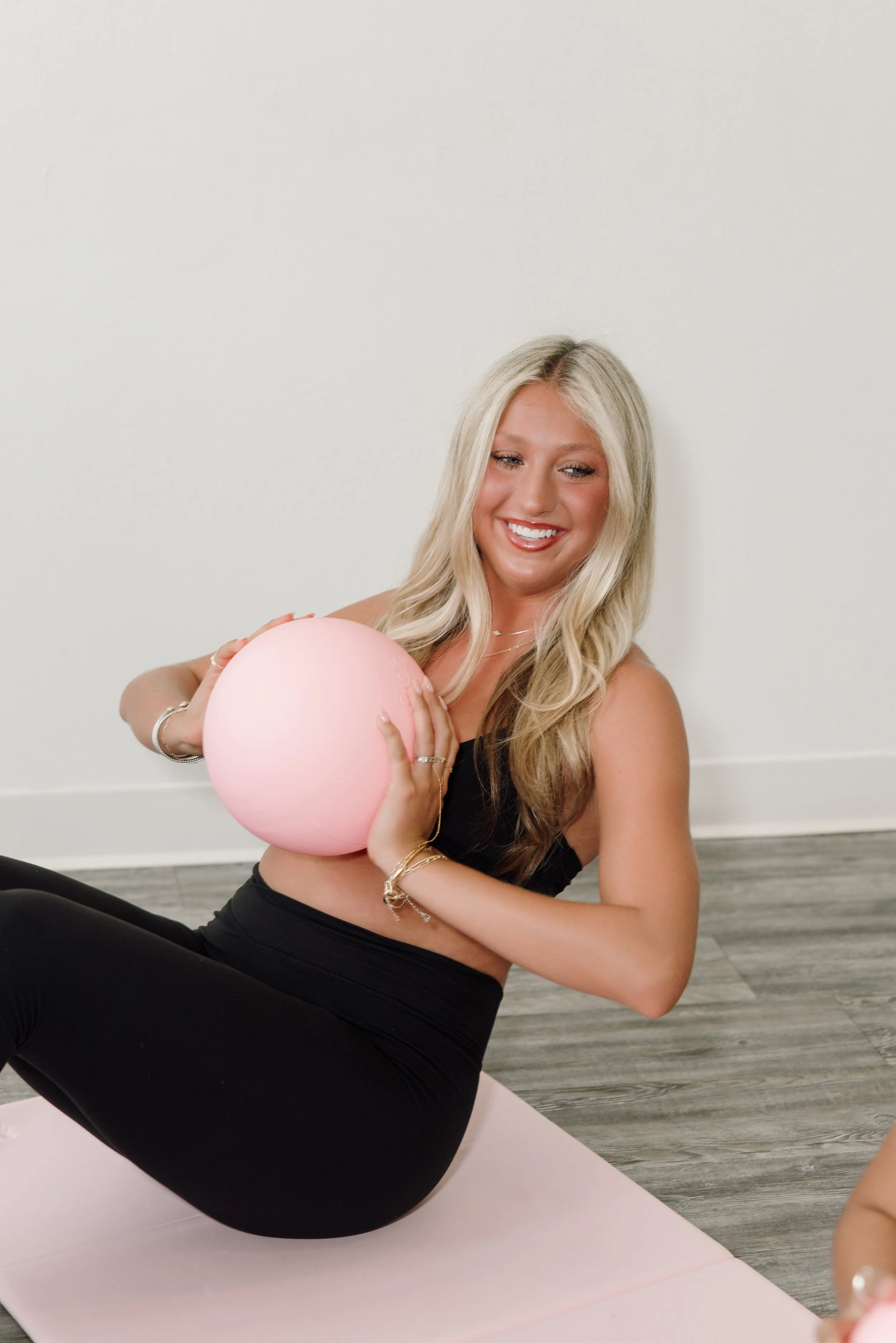 Smiling young woman with long blonde hair holding a pink balloon while sitting on a pink yoga mat, wearing a black sports bra and black leggings, in a room with grey wooden floors and a plain white wall.