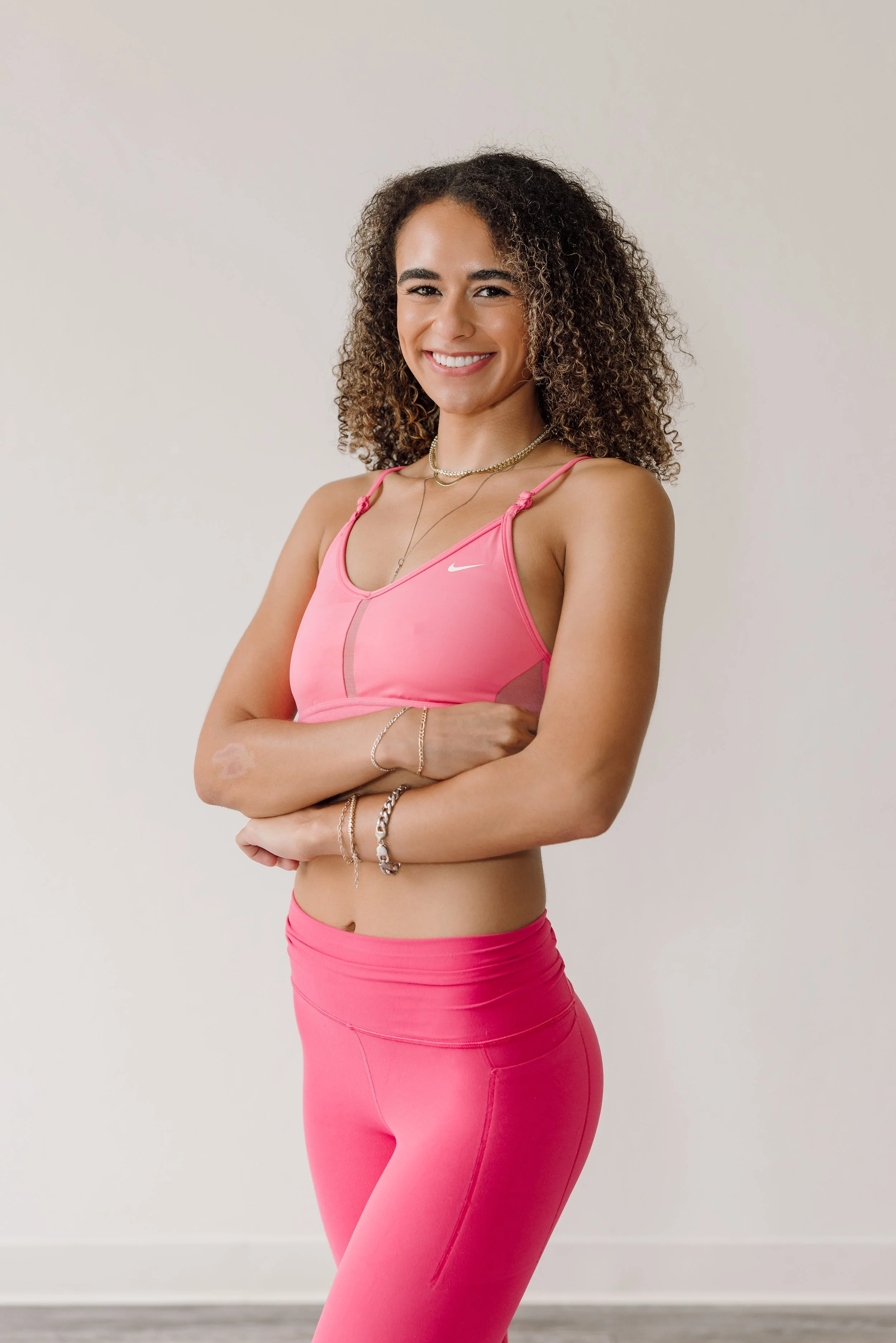 A young woman with curly brown hair smiling and standing with arms crossed, wearing a pink sports bra and matching pink leggings, accessorized with layered necklaces and bracelets, against a plain off-white background.