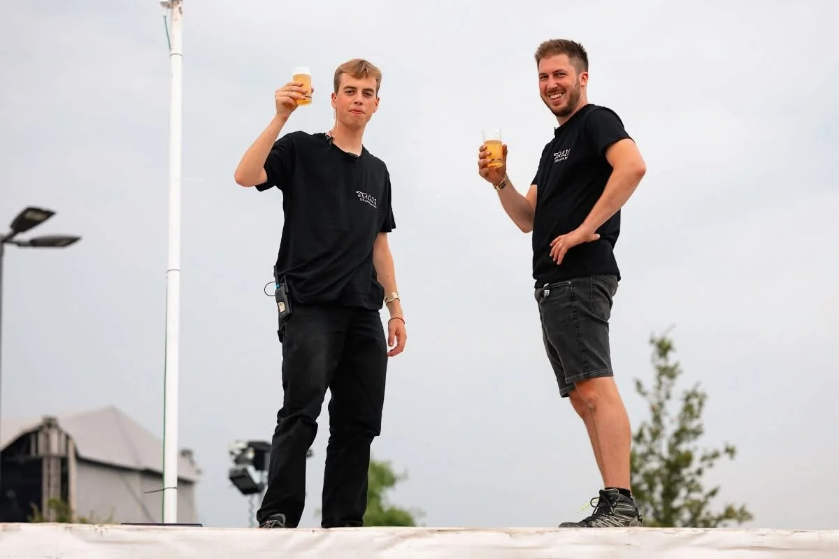 een jongen met lichtbruin haar en een zwarte T-shirt met de tekst 'TRAX FESTIV ORGANISATIE' staat binnen in een ruimte met een scherm op de achtergrond.
