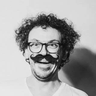 Portret van een man met curly haar, een bril en een grote snor, lachend