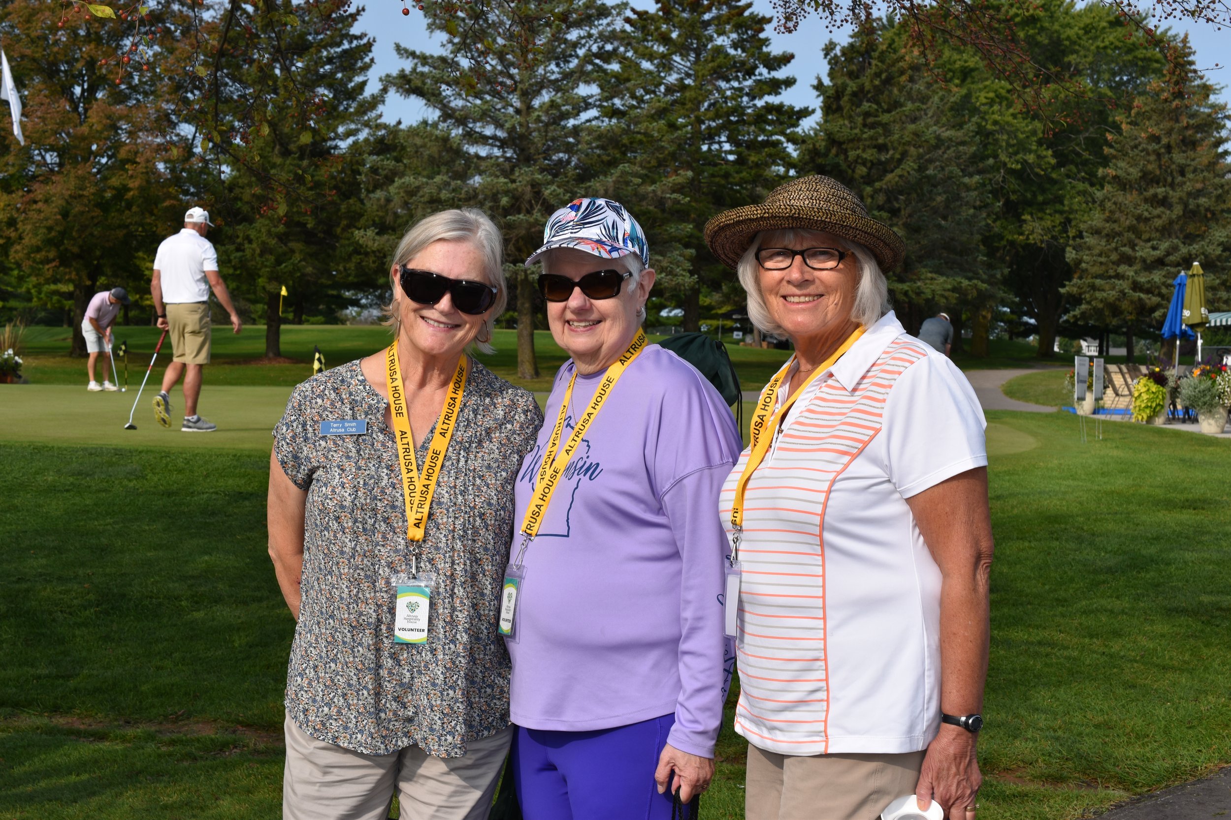Three women wearing name tags and lanyards smiling on a golf course with golfers in the background.