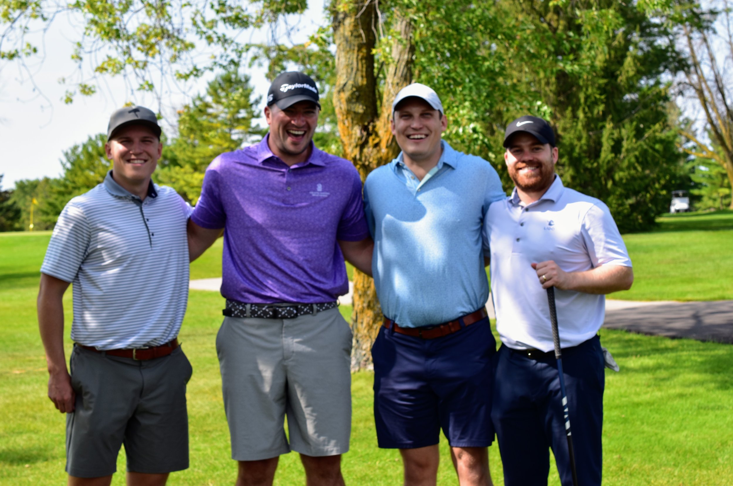 Four men posing on a golf course, smiling, wearing caps and golf attire.