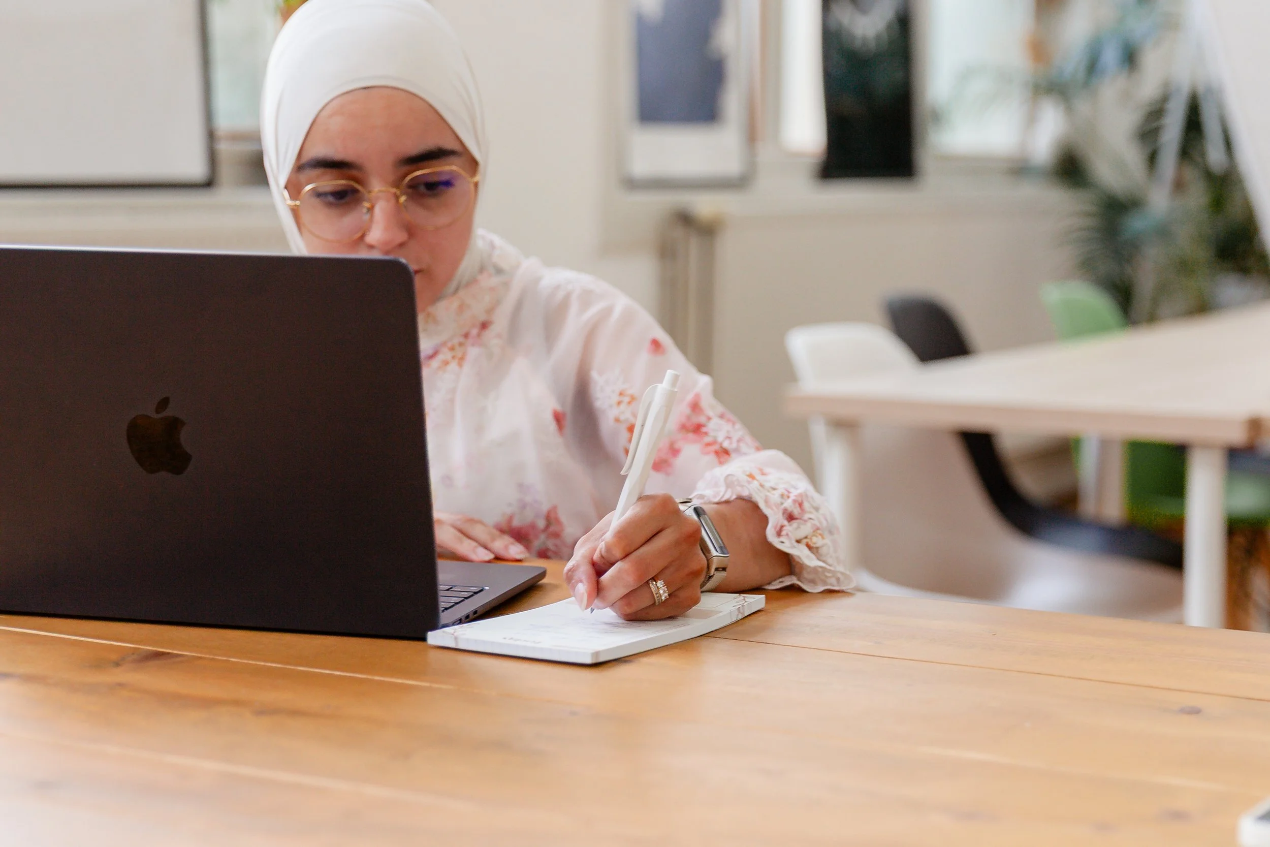 Vrouw met hoofddoek werkt aan een houten tafel met een laptop, notitieboekje en pen in een moderne, lichte kantoorruimte.