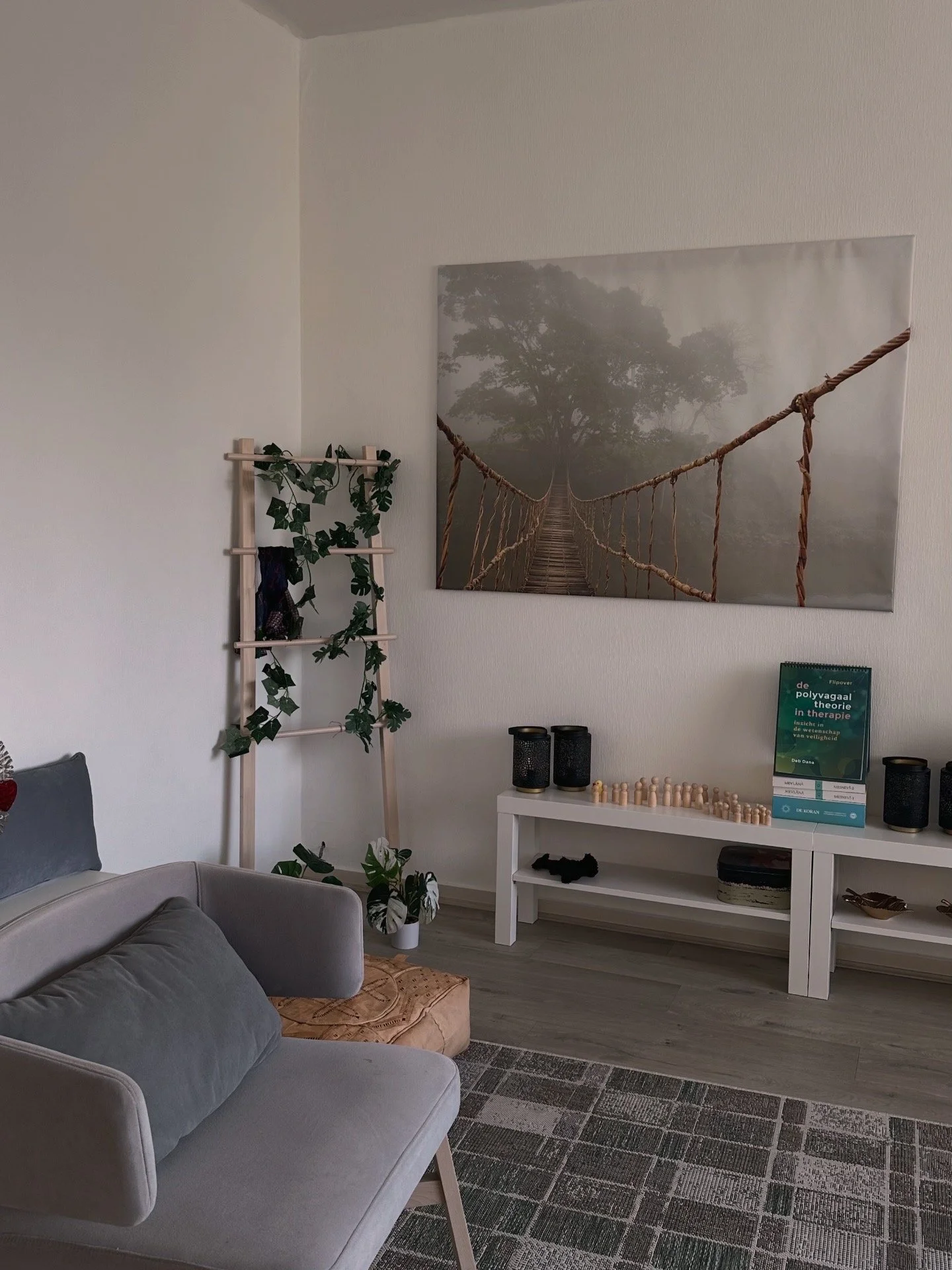 Een woonkamer met een beige bank, een witte salontafel met schoorstenen en een boek, een boom- en klimrek met planten, een grote muurkunst van een touwbrug in de jungle, en een grijs en bruin tapijt op de vloer.