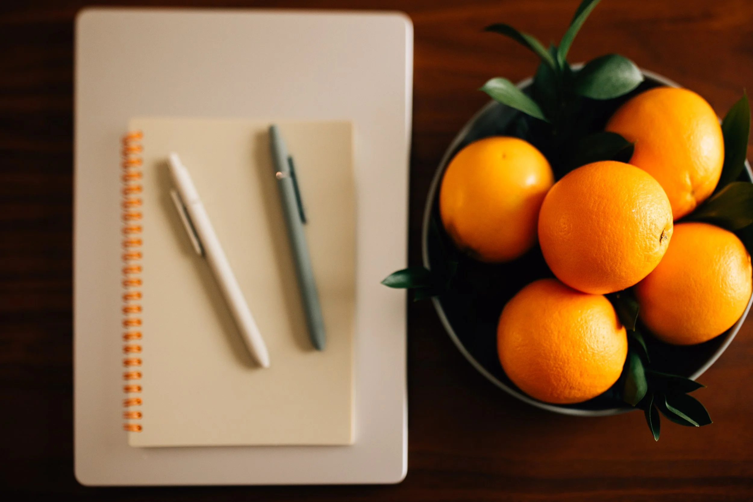 A top view of a notebook with two pens on it and a bowl of six oranges with green leaves, on a wooden surface.