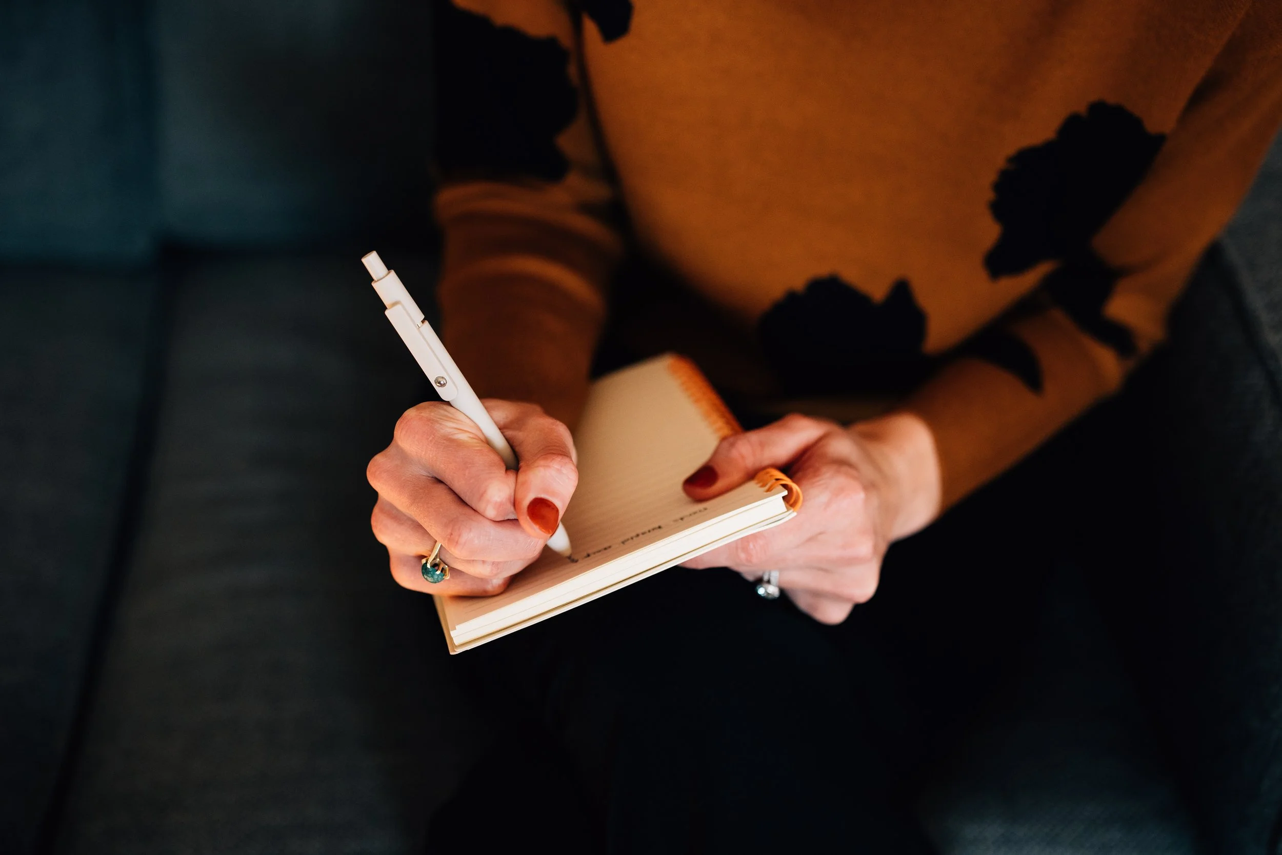 Person writing in a notebook with a white pen, sitting on a dark-colored sofa